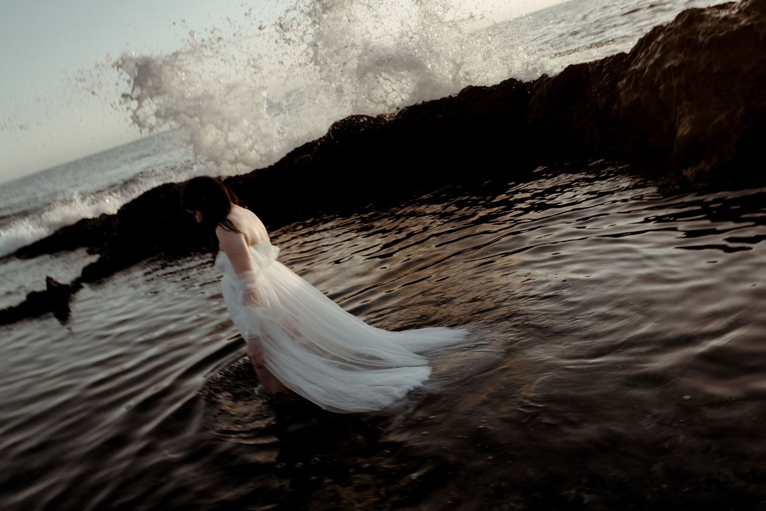A flowing white wedding dress floats gracefully in dark ocean waters during an underwater photography session.