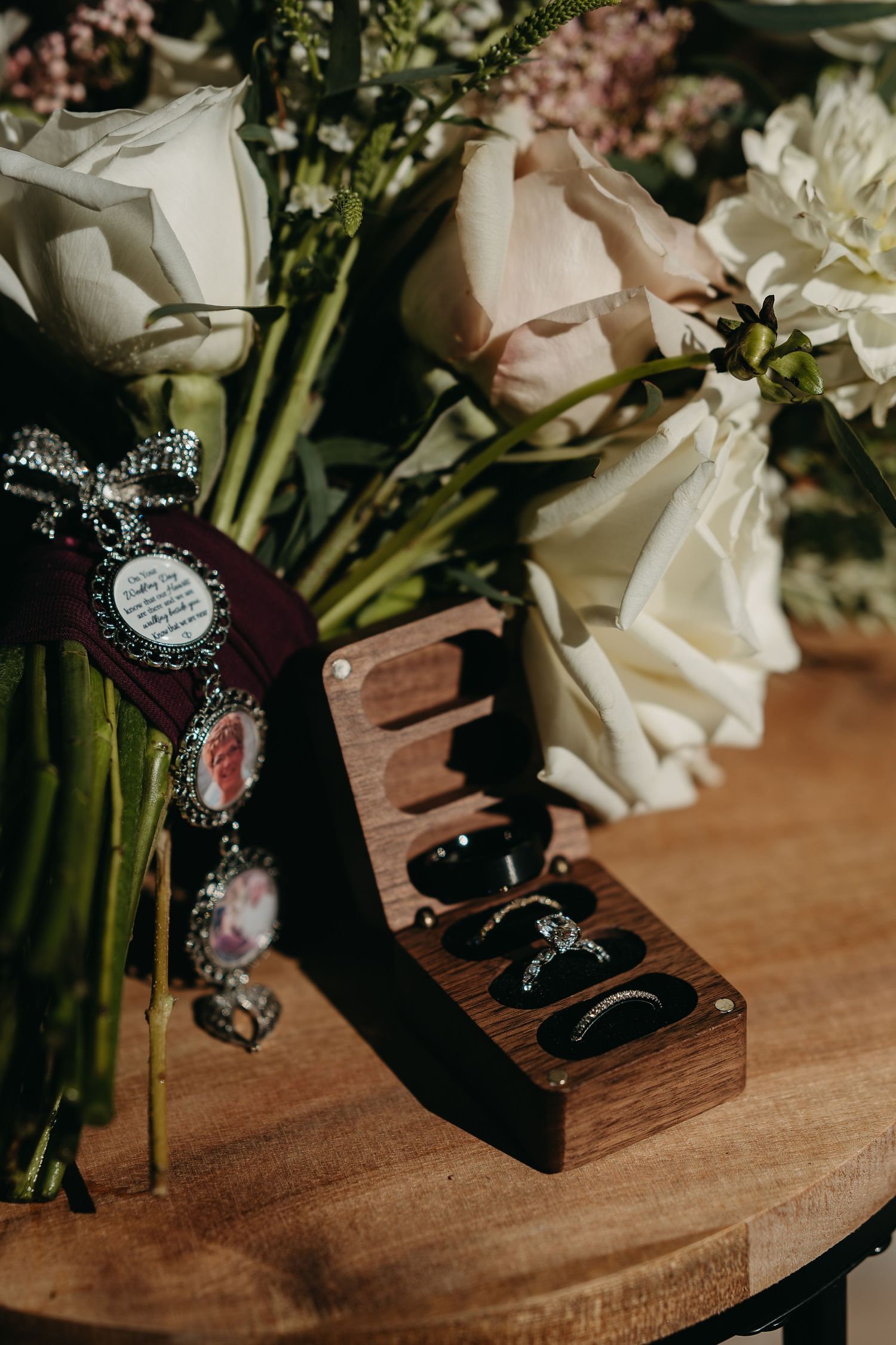 A wooden jewelry display box with rings sits beside pink and white flowers on a round wooden table.