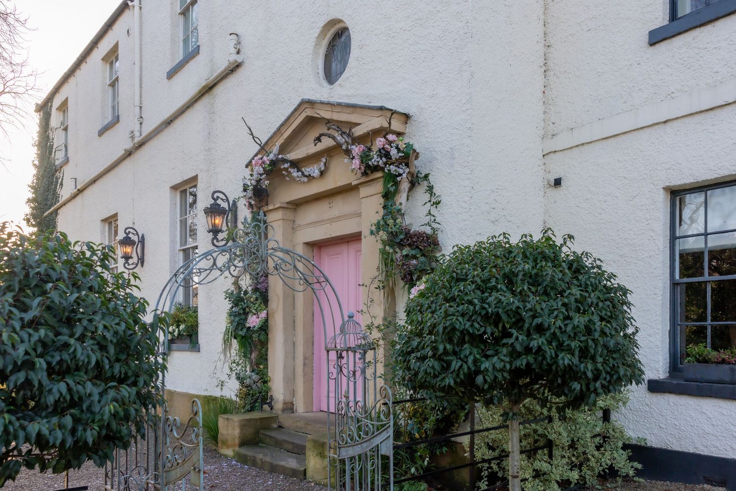 Elegant pink door with ornate stone archway and decorative greenery on white building facade.