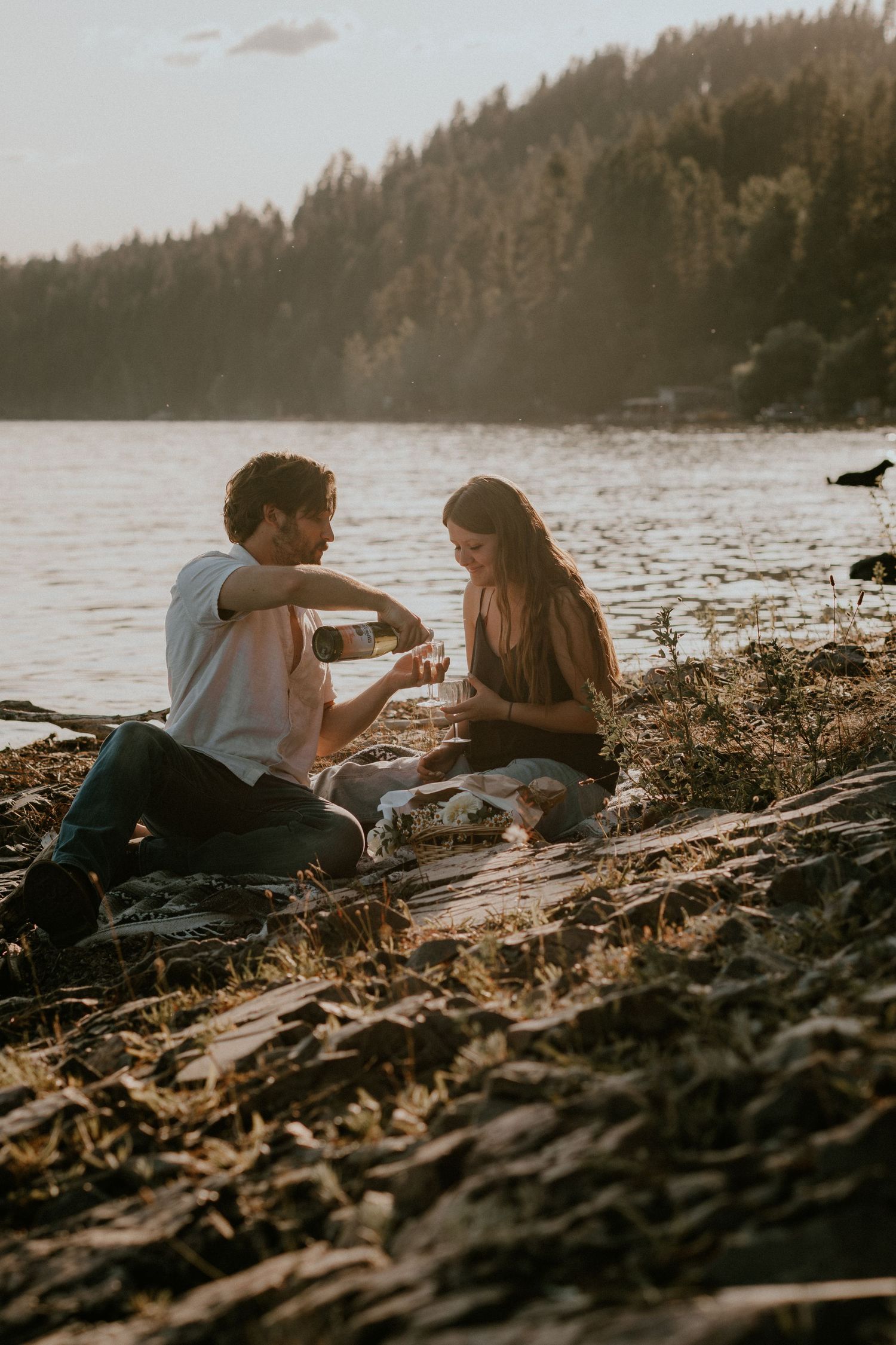 A couple enjoys wine at sunset on a rocky lakeshore with evergreen trees in the background.