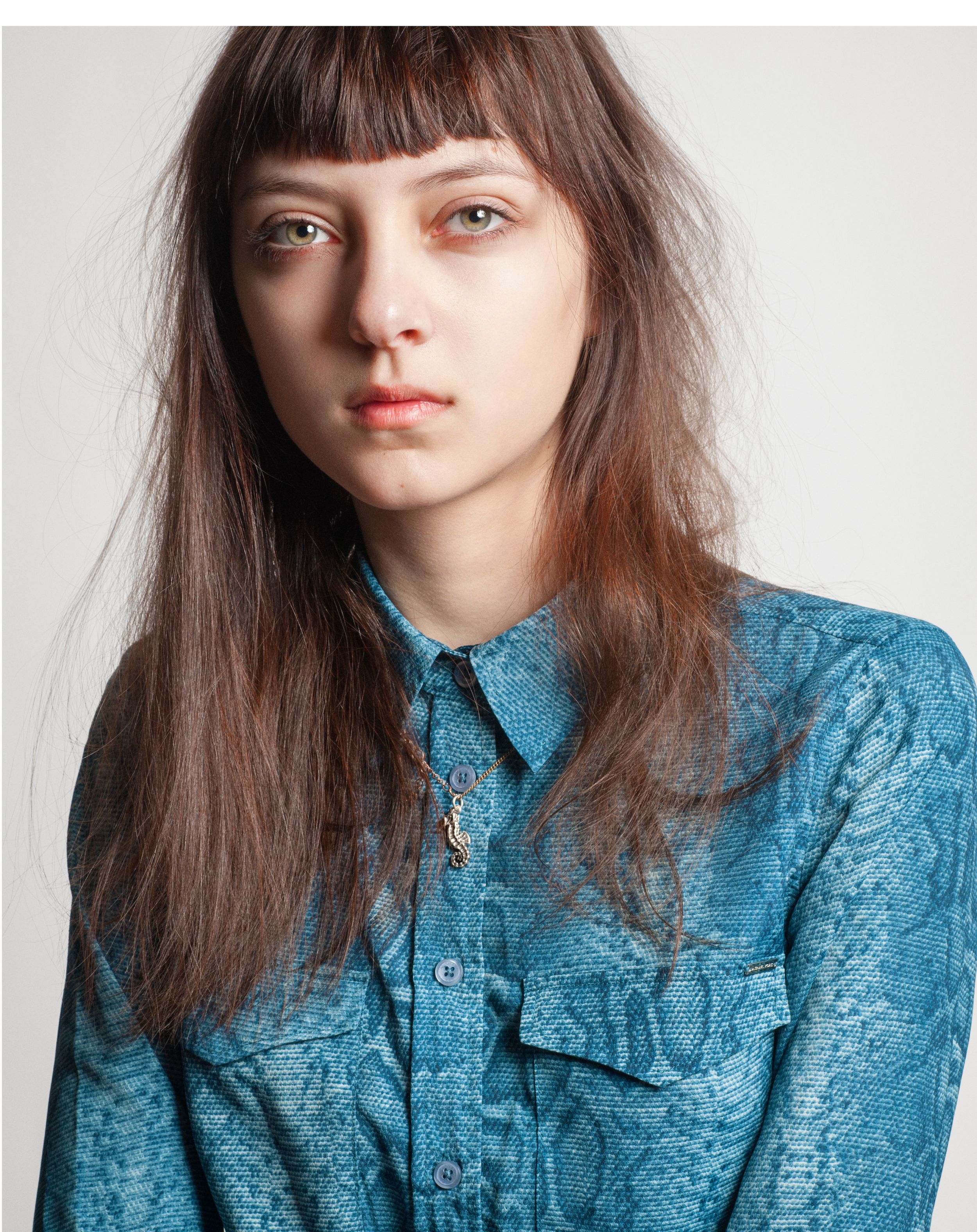 Young woman with brown hair and bangs wearing a teal snake print button-up shirt, posing for a portrait photo.
