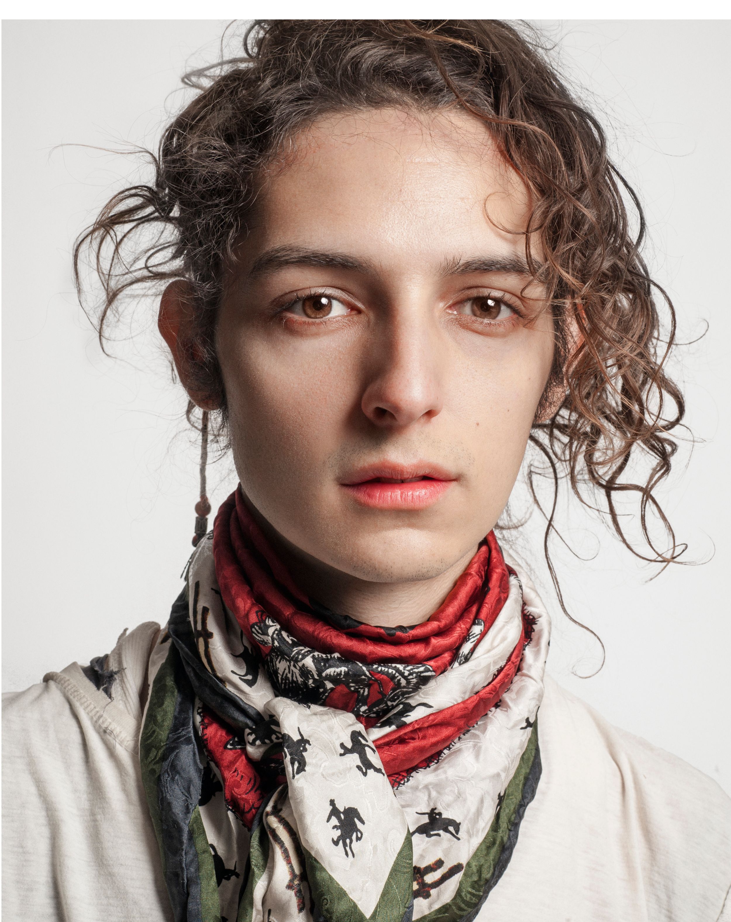 Young man with curly brown hair wearing a patterned red scarf and white jacket against a white background.
