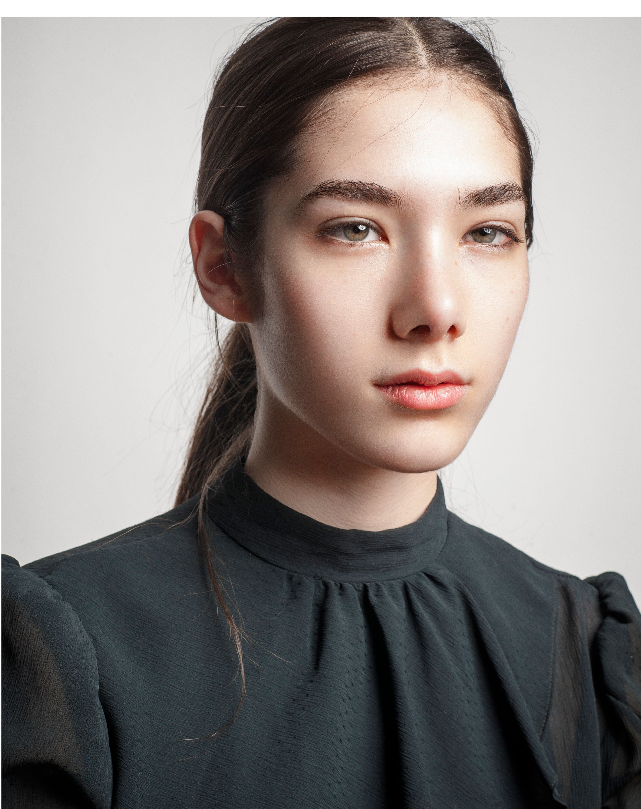 Young woman with dark hair in ponytail wearing black high-neck puff sleeve top, posing against gray background.