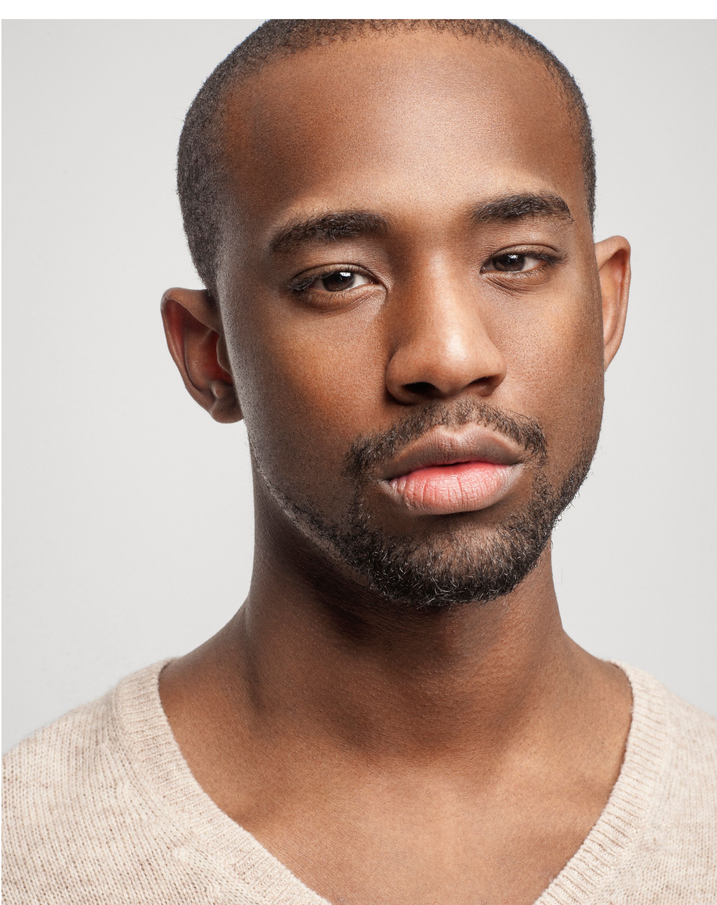 Young Black man with short hair and beard wearing a beige v-neck sweater against a light gray background.