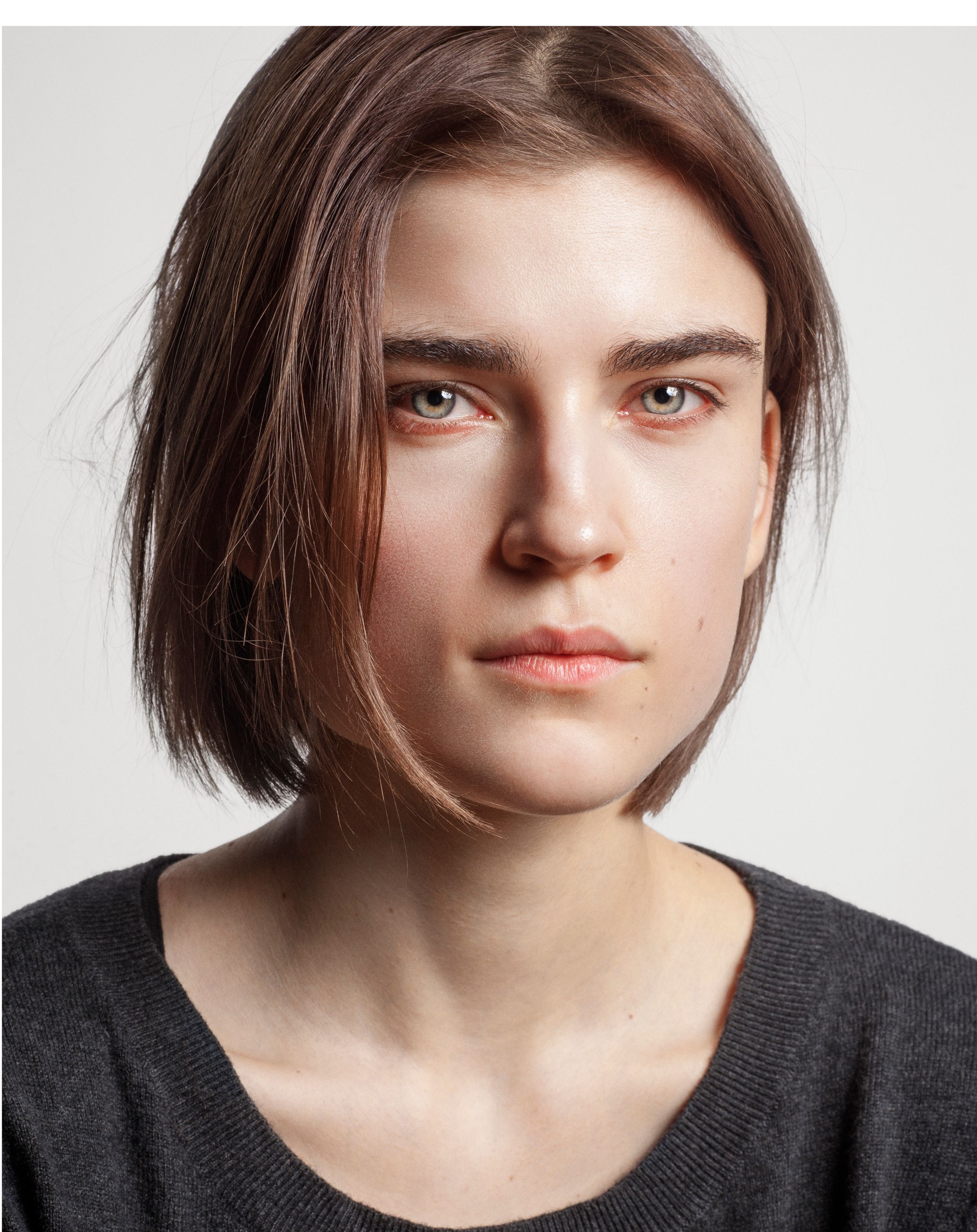 Young woman with short brown bob haircut wearing a dark gray top against a white background.