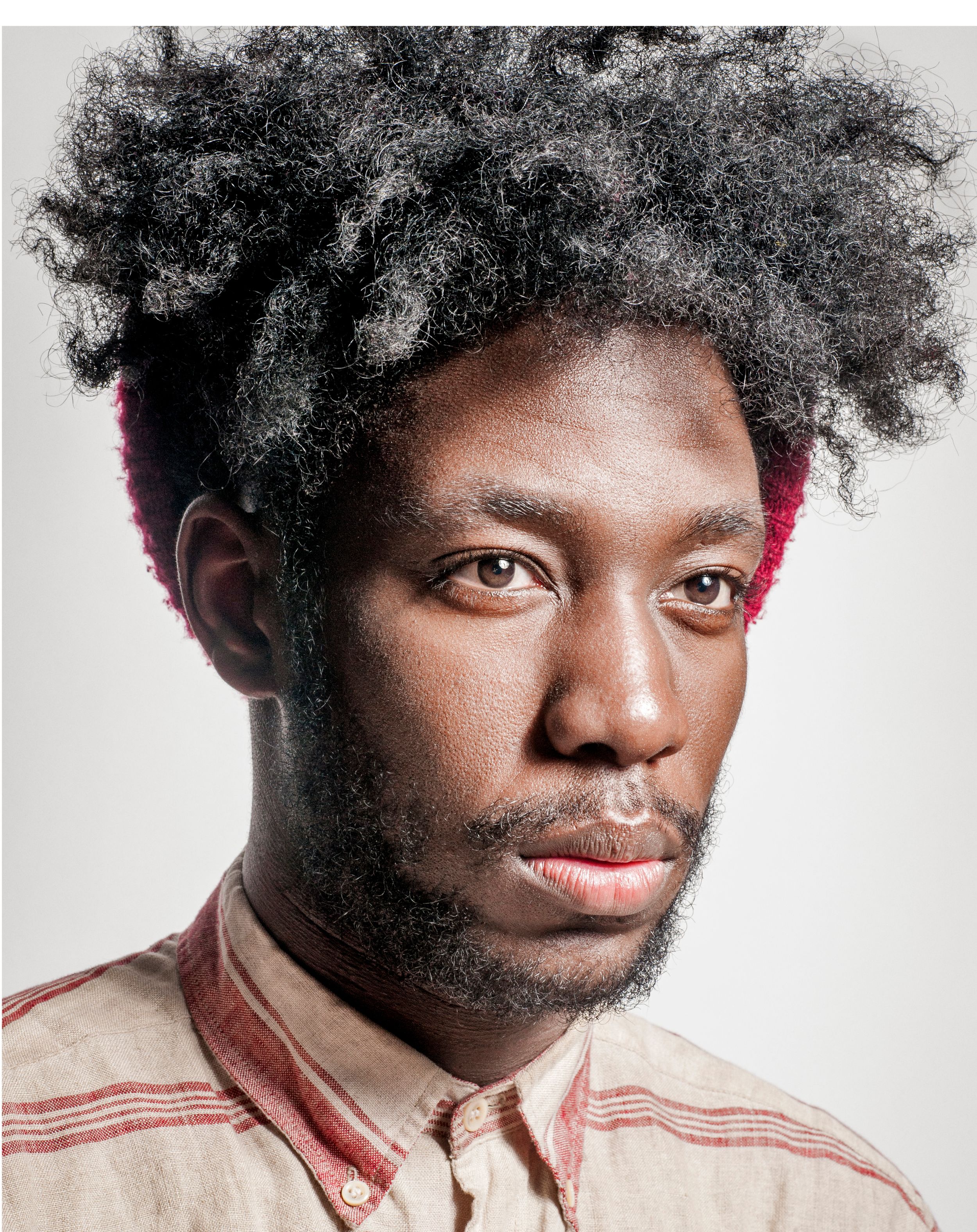 Young Black man with natural afro hair, light beard, wearing a striped linen shirt, posing against a gray background.