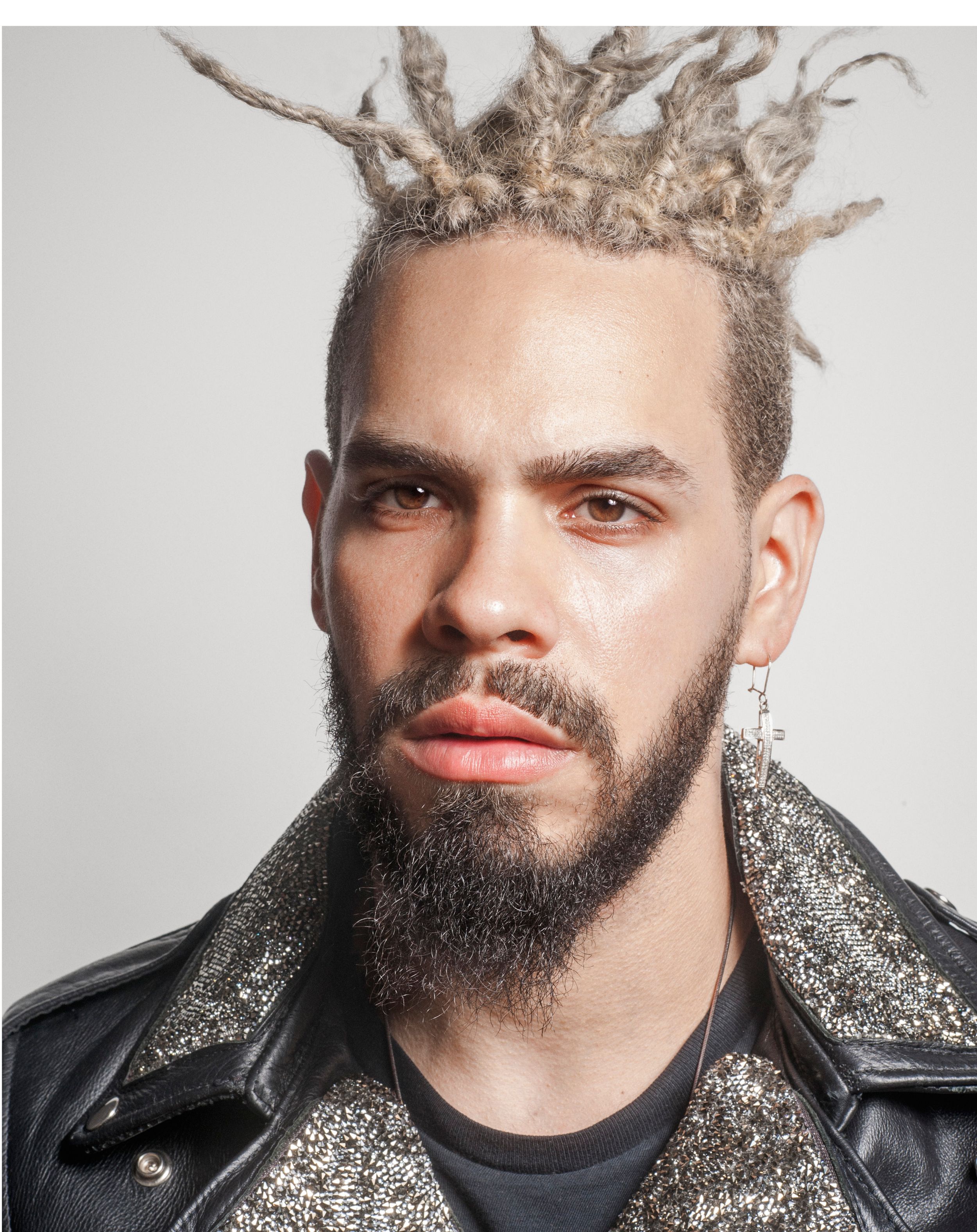 Man with bleached dreadlocks, dark beard, earring, wearing a glittery jacket and black leather coat.