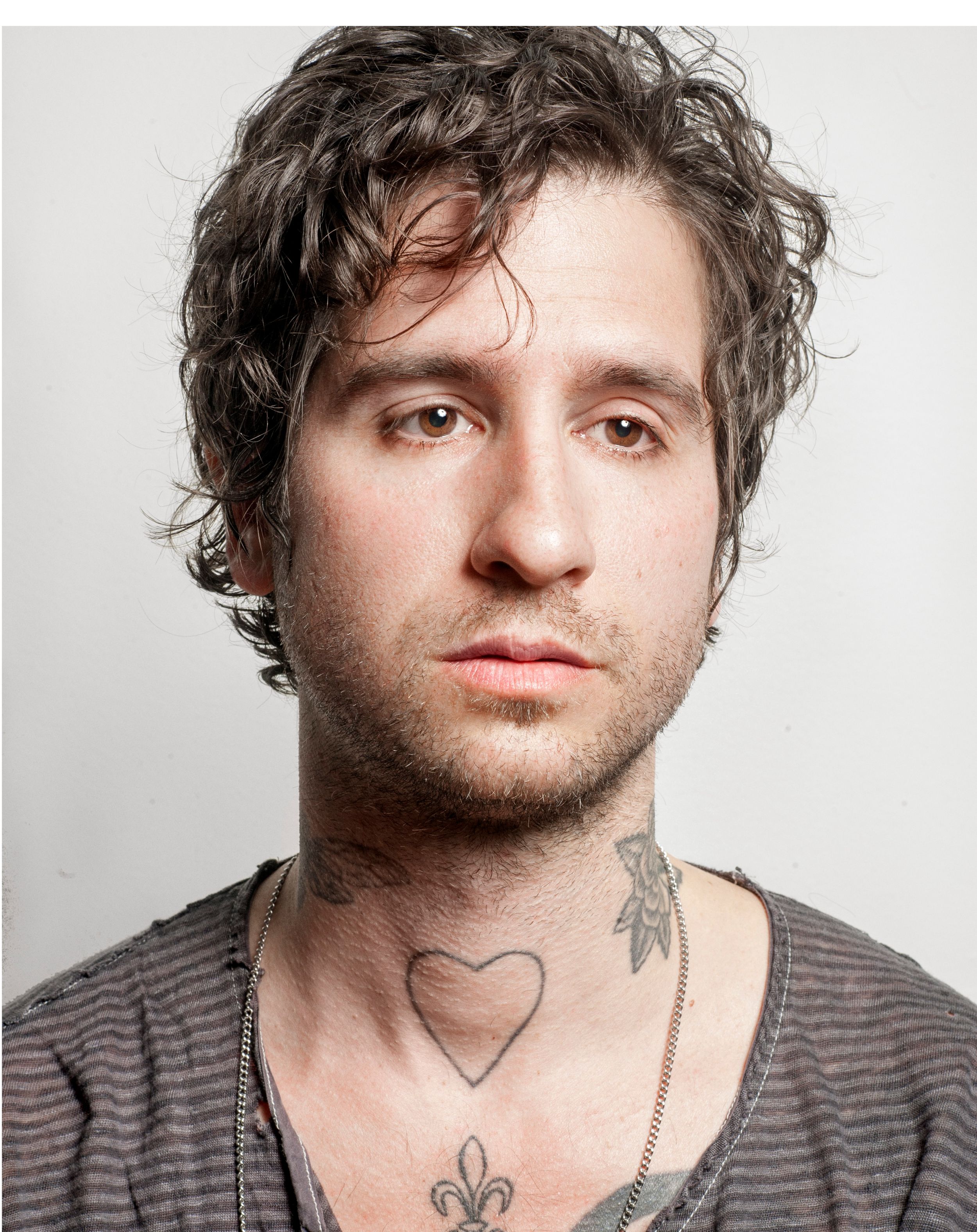 Young man with curly dark hair, heart and fleur-de-lis chest tattoos, wearing a striped shirt and chain necklace.