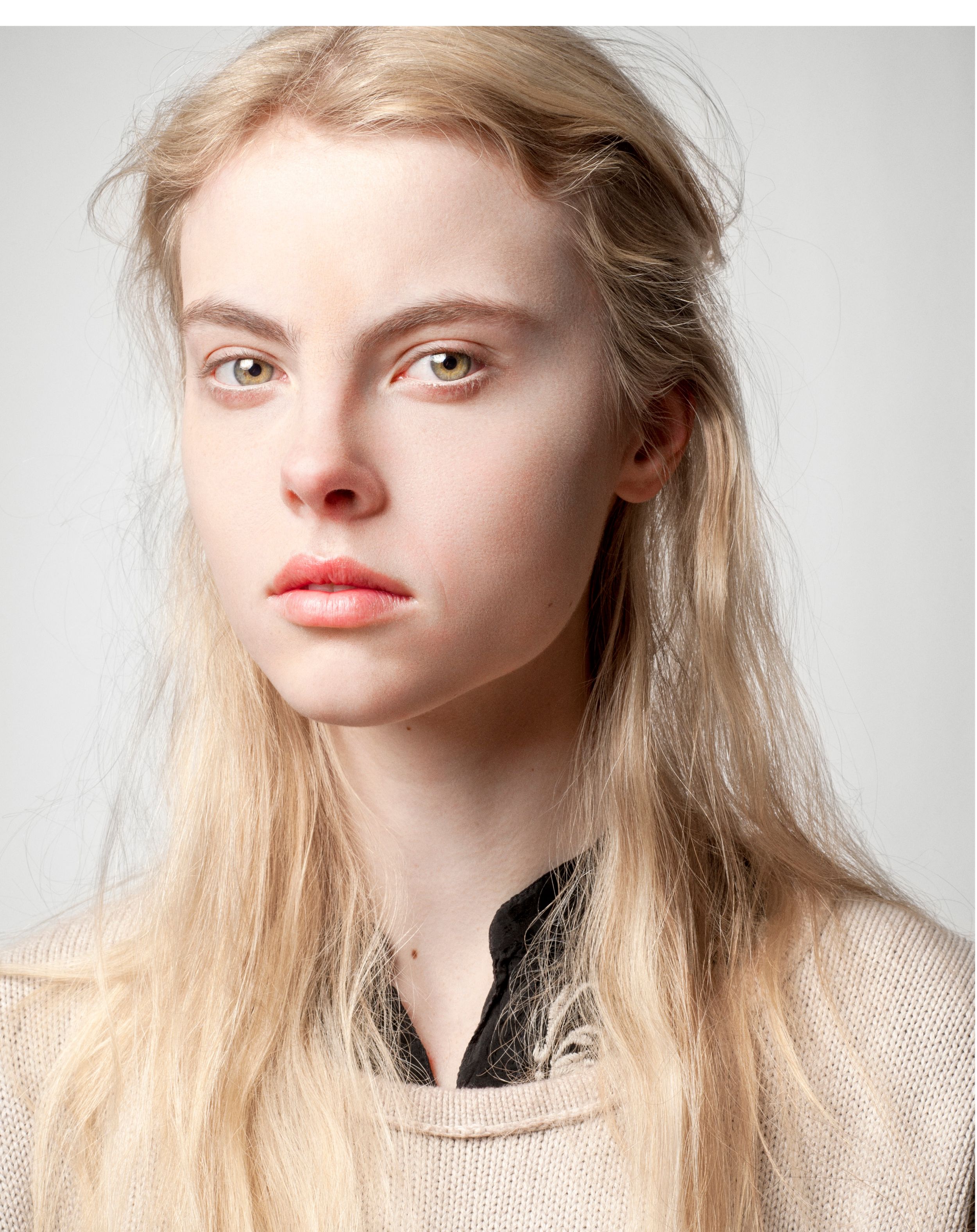 Young woman with long blonde hair and minimal makeup wearing a beige knit top against a light gray background.