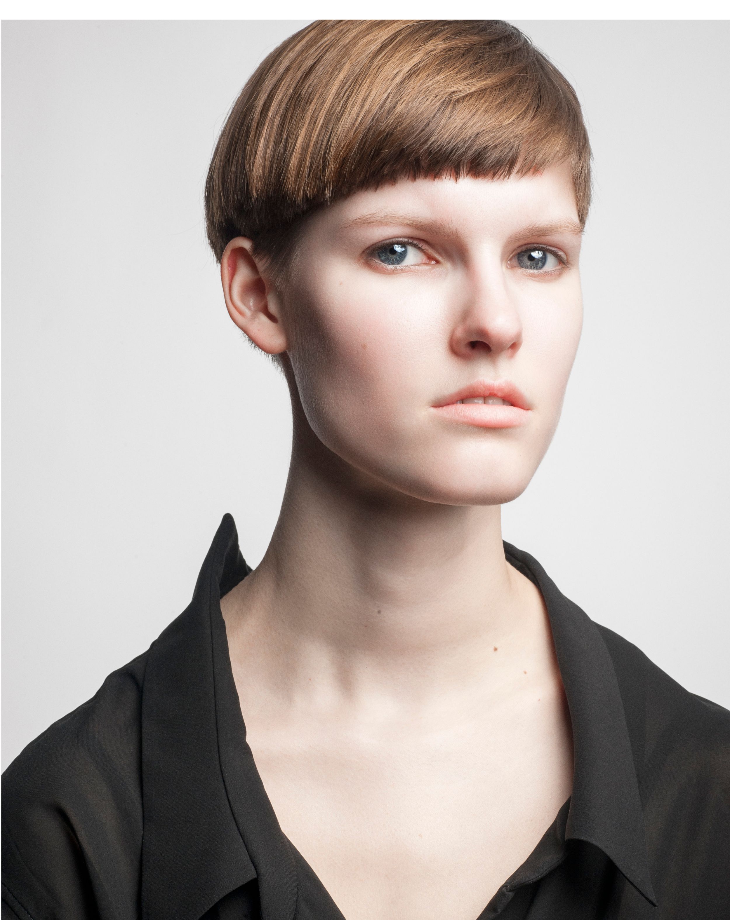 Young woman with short brown bowl cut hair wearing a black shirt, photographed against a light gray background.