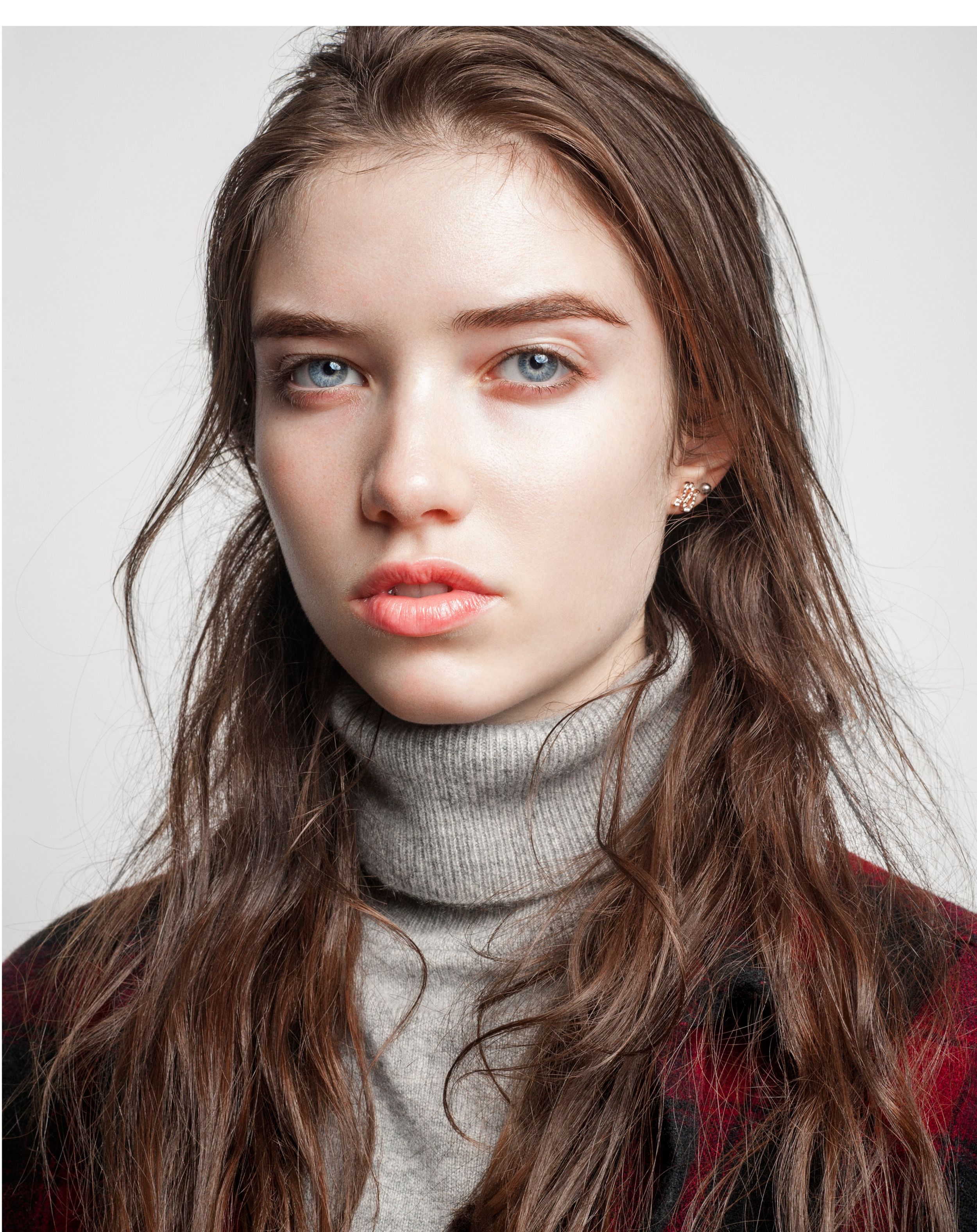 Young woman with blue eyes and long brown hair wearing a gray turtleneck sweater and colorful jacket.