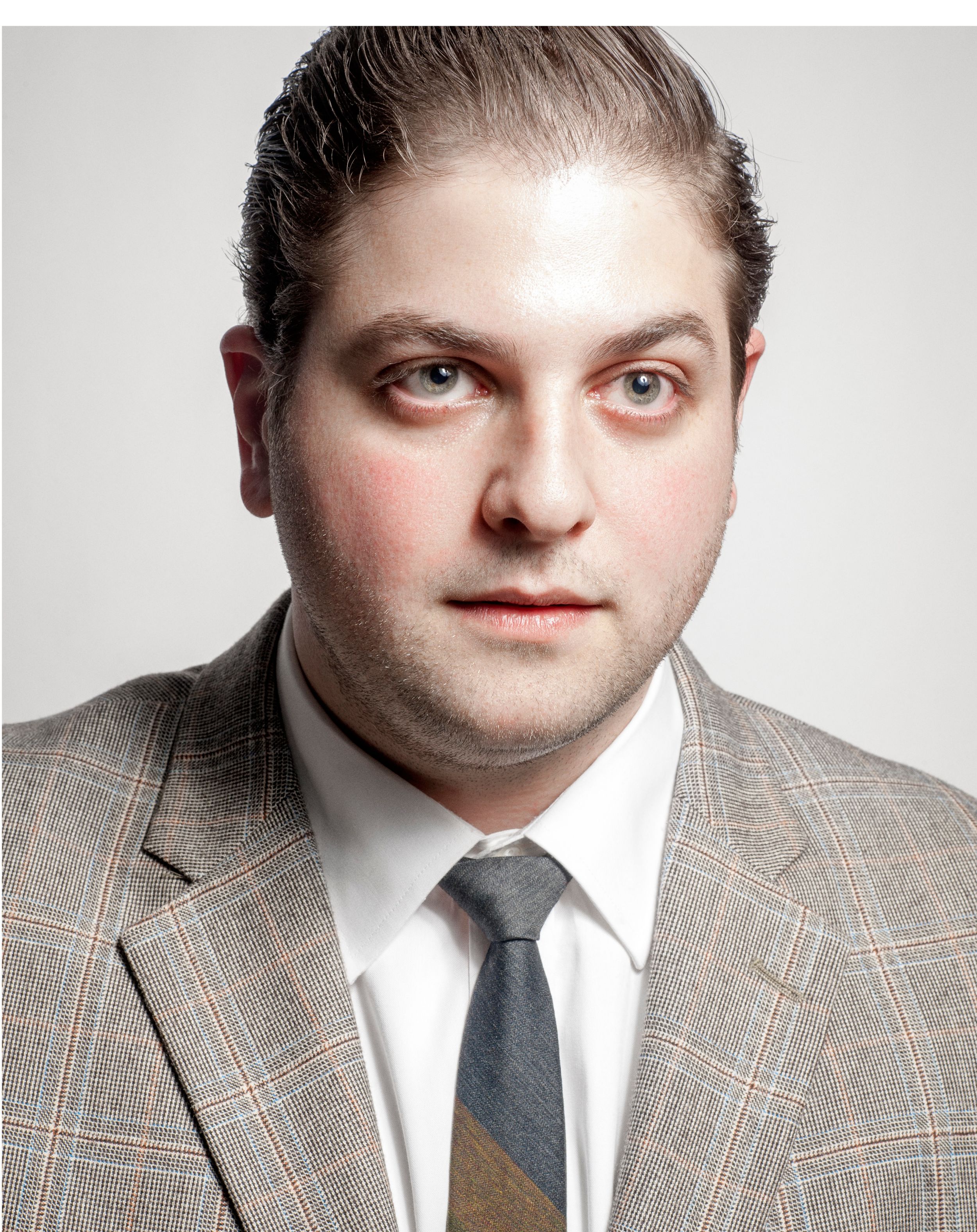 Professional headshot of a man in a gray plaid blazer and striped tie against a neutral background.
