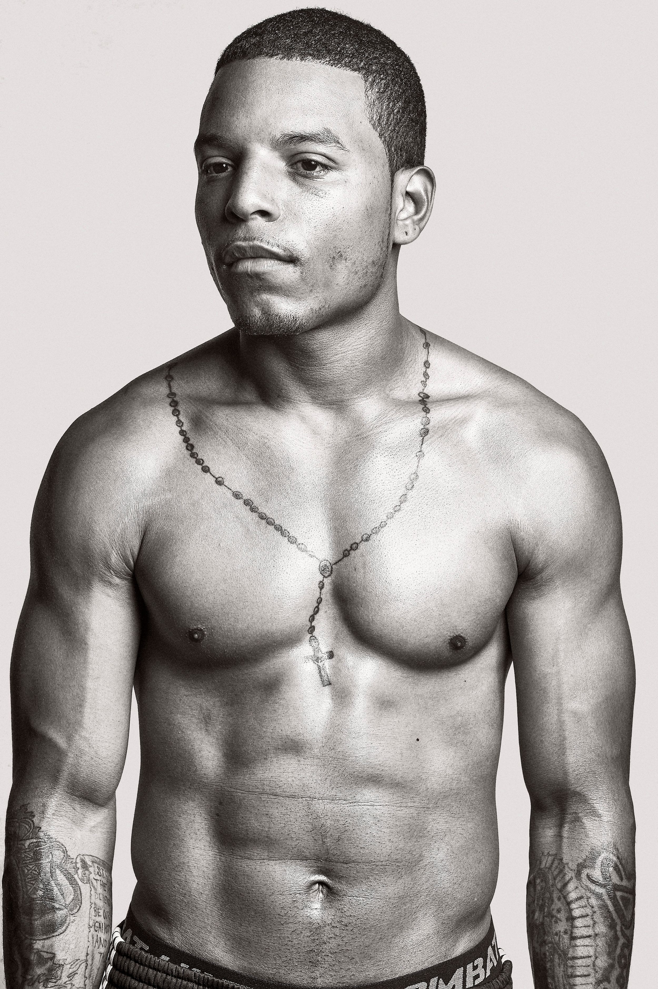 Muscular shirtless man with tattoos and cross necklace poses confidently in black and white portrait photo.