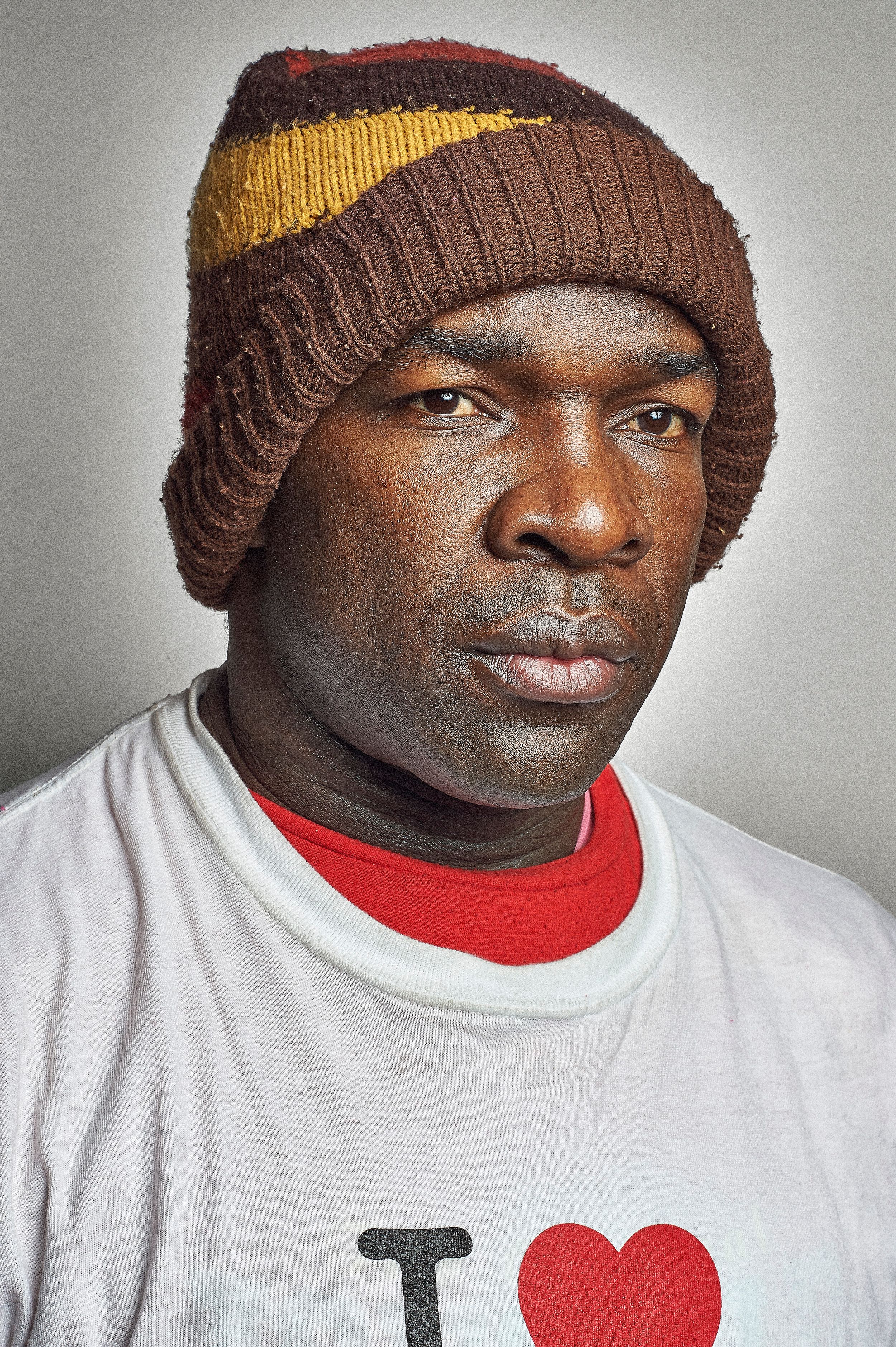 Man wearing a brown, red, and yellow knit beanie hat and a white I Love graphic t-shirt against a gray background.