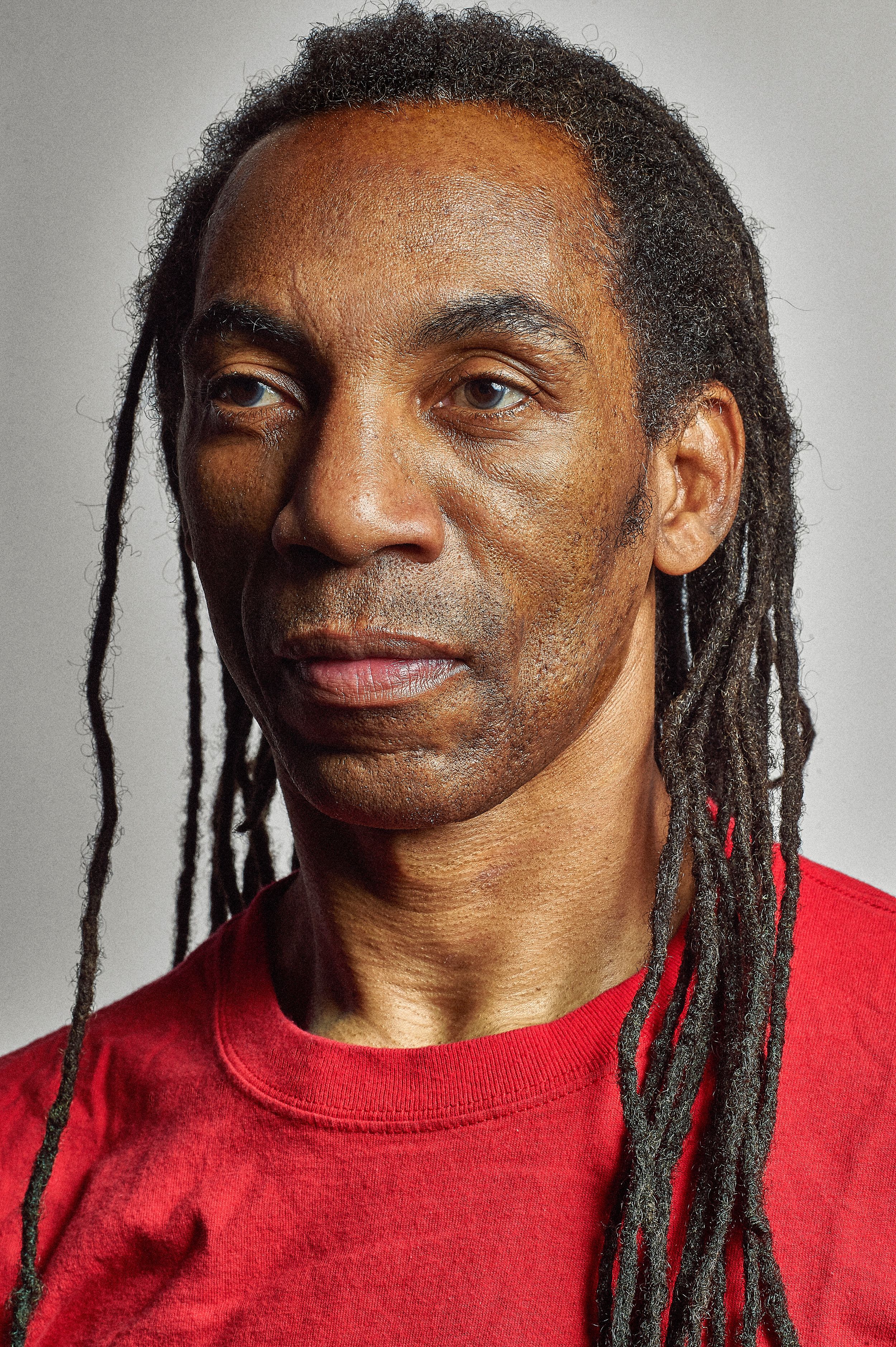 Middle-aged Black man with long dreadlocks wearing a red t-shirt, photographed against a gray background.