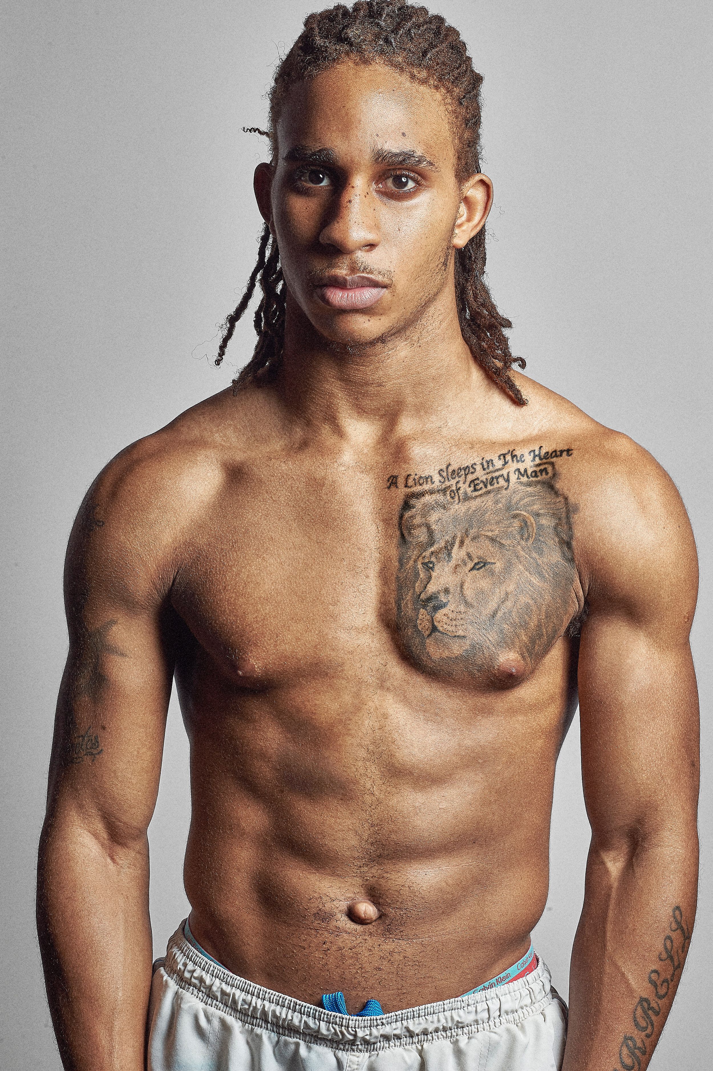 Muscular shirtless man with braids and a lion chest tattoo posing against a gray background.