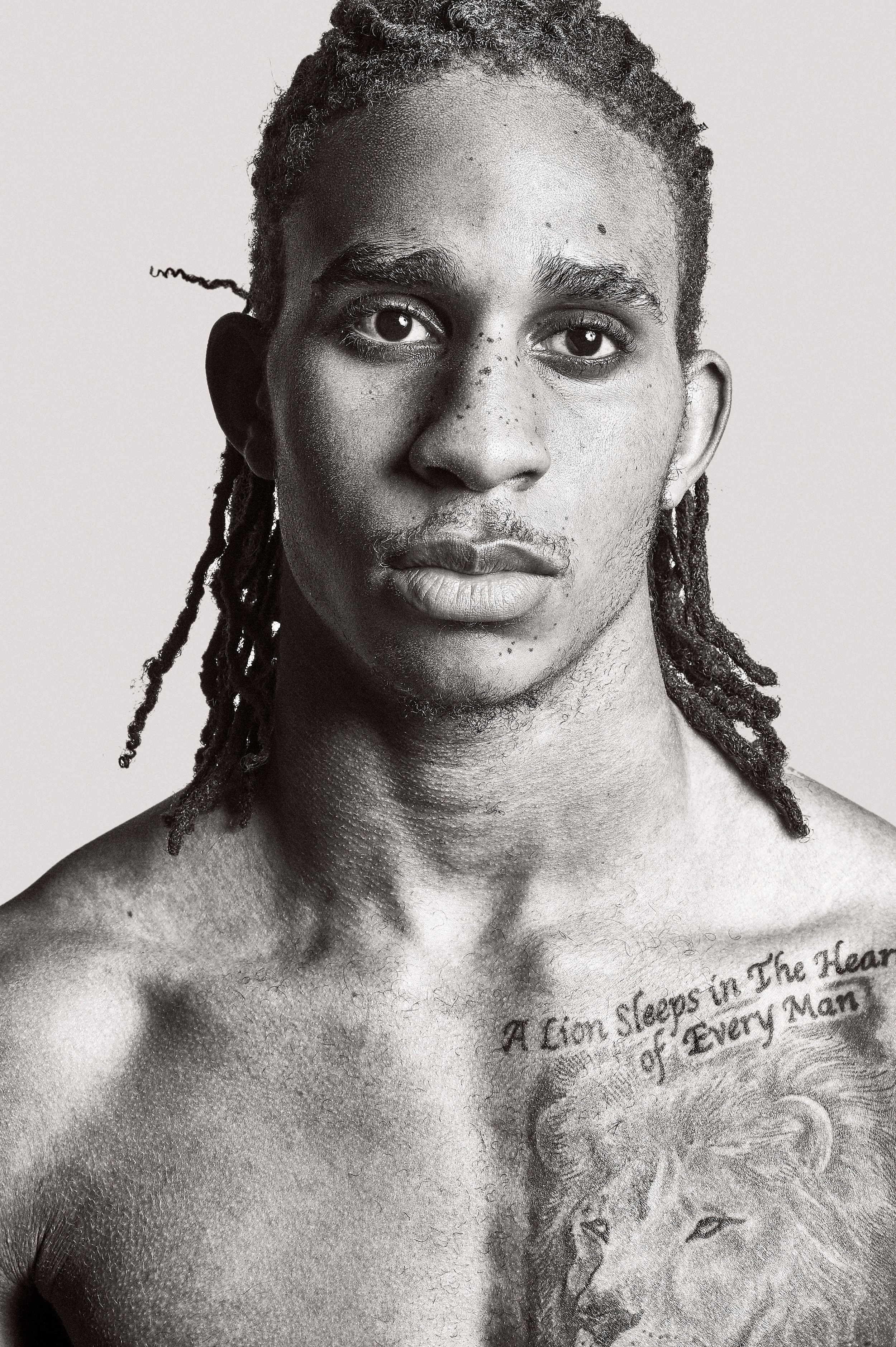 Black and white portrait of a young man with dreadlocks and a lion chest tattoo reading A Lion Sleeps in the Heart of Every Man.