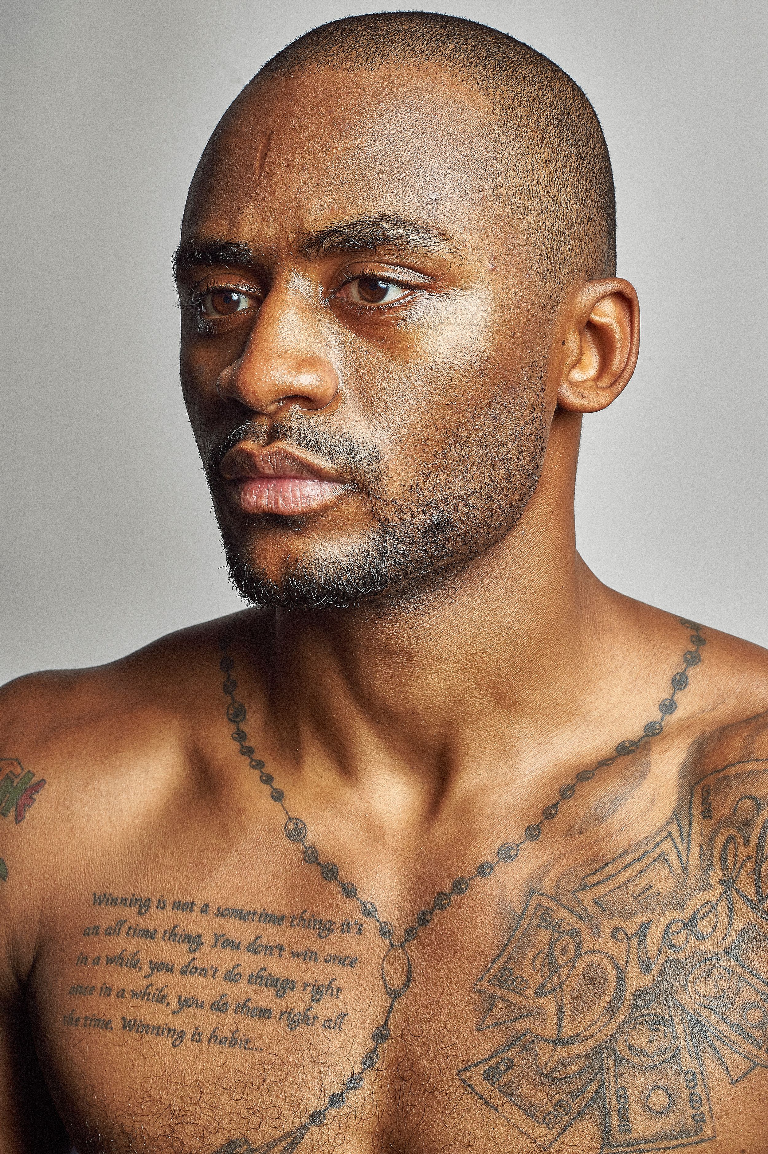 Shirtless man with tattoos on chest and shoulders posing against gray background.