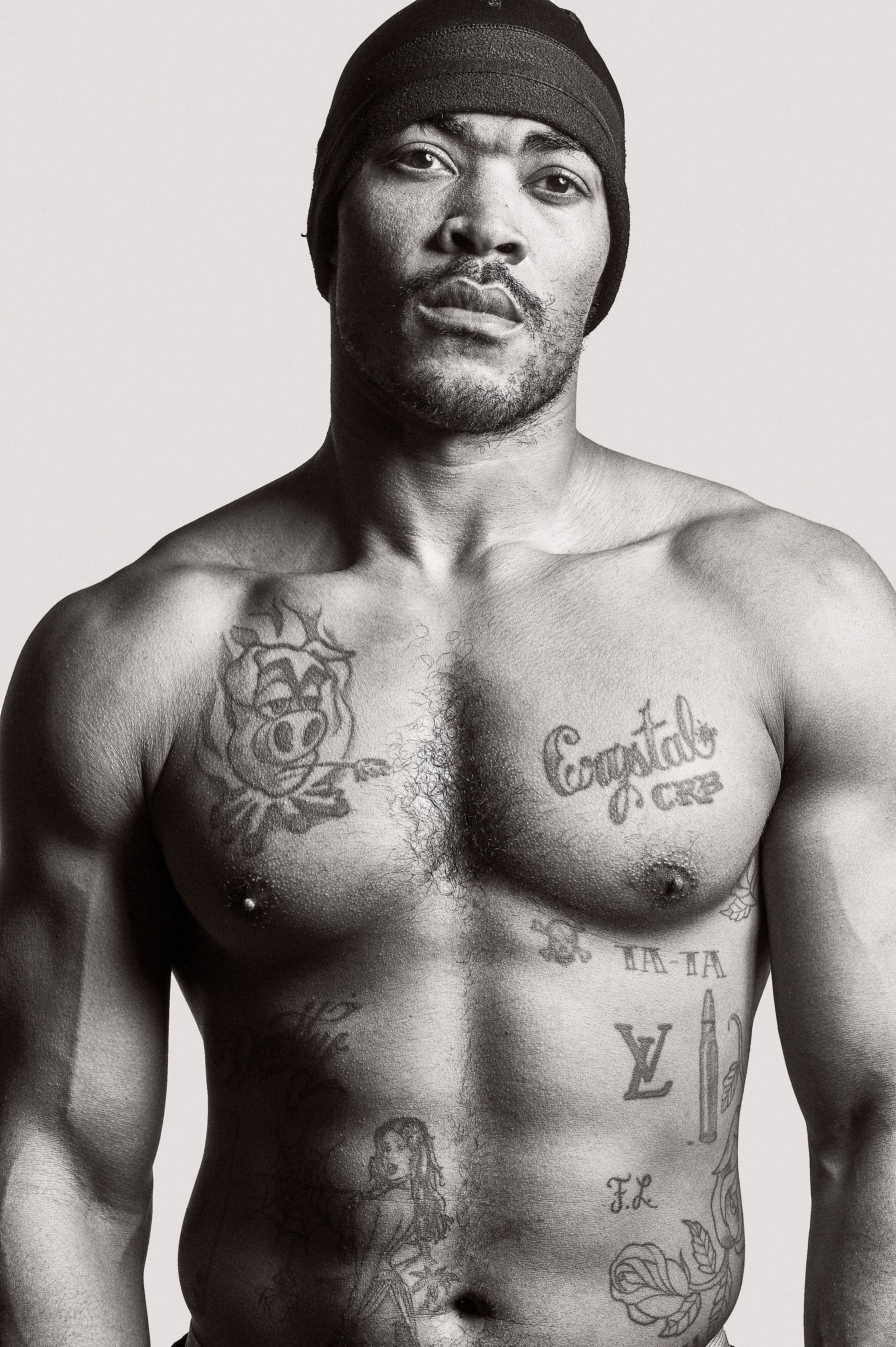 Black and white photo of a muscular shirtless man with tattoos wearing a black beanie hat.