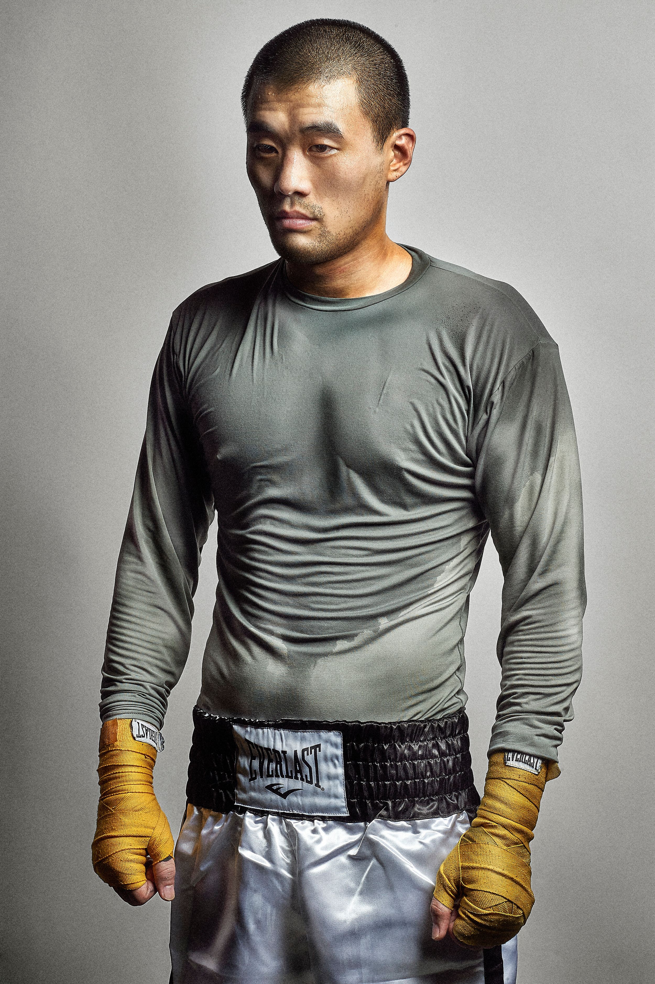 Male boxer in gray long-sleeve shirt and silver Everlast shorts with gold hand wraps, posing against gray background.