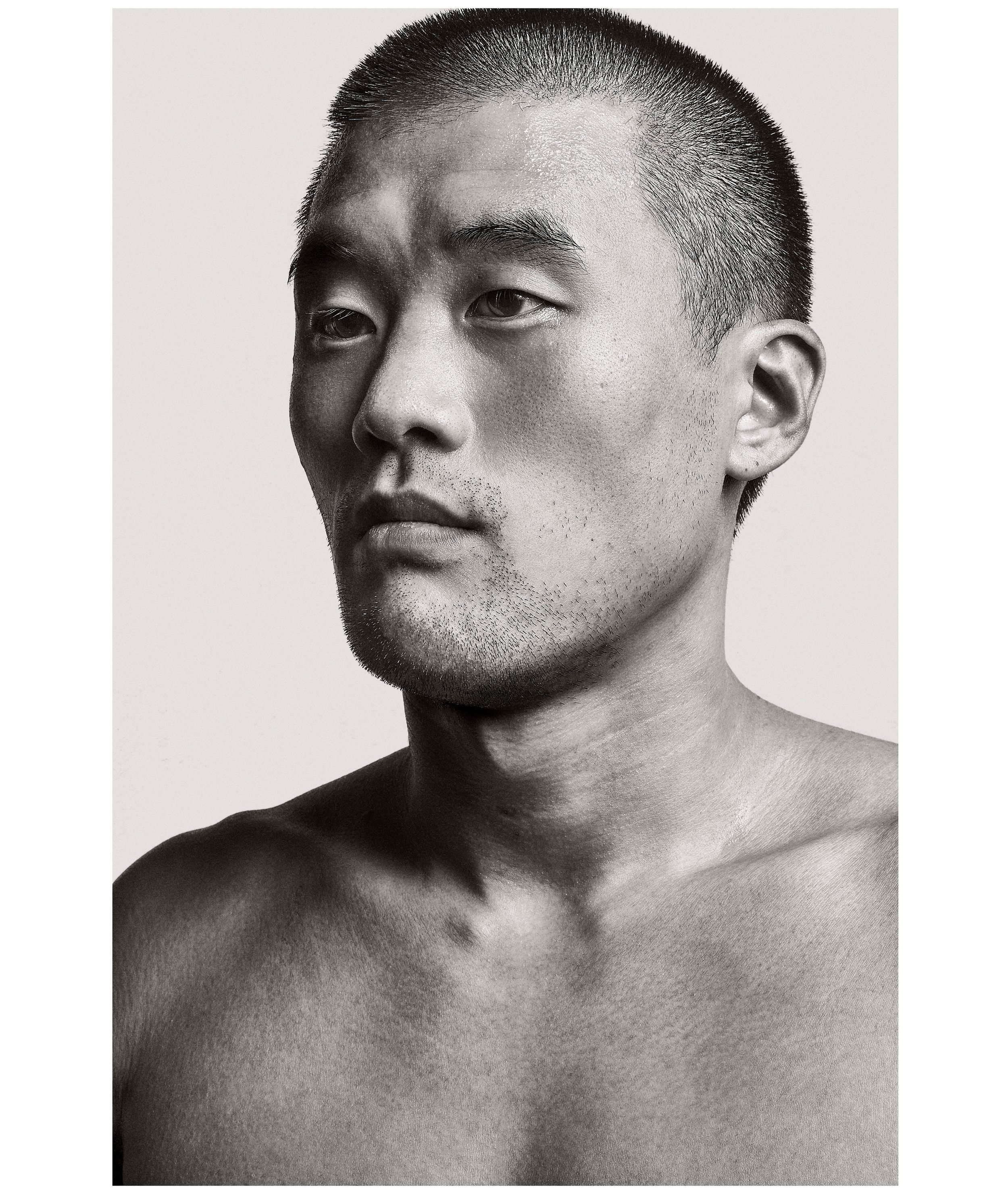 Black and white portrait of a shirtless young Asian man with short hair against a light background.