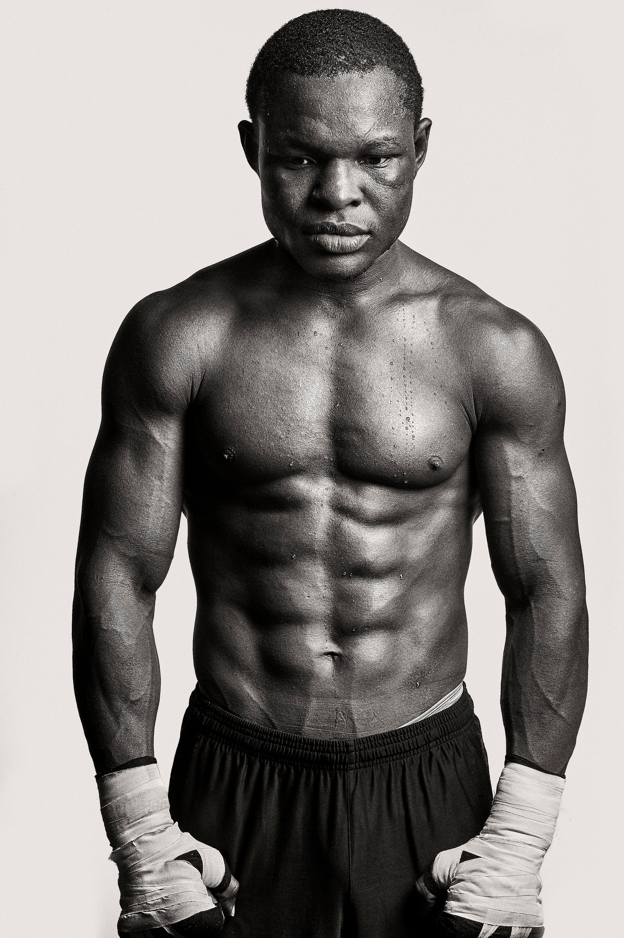 Black and white photo of a muscular shirtless male boxer with hand wraps, showcasing defined abs and athletic physique.
