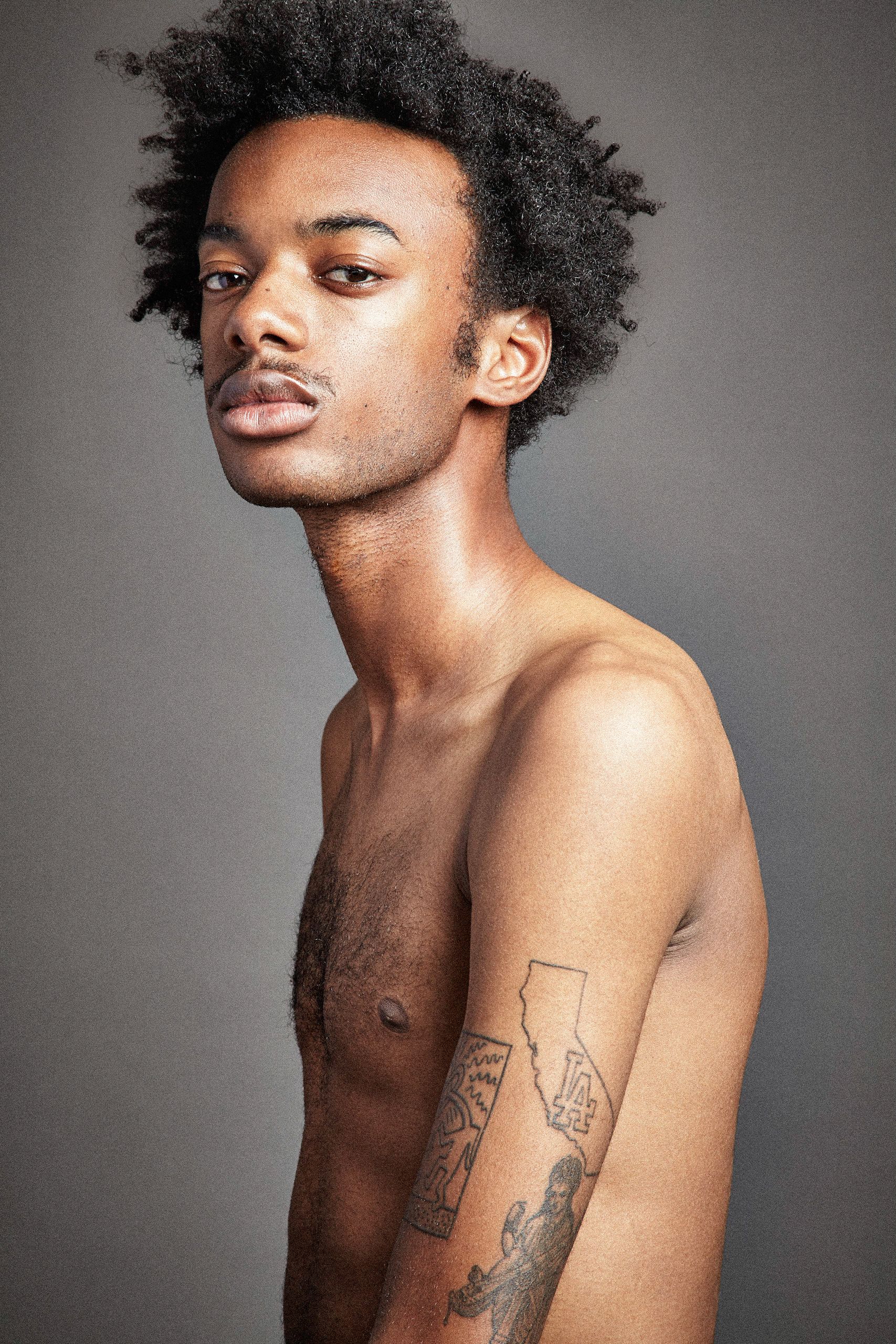 Studio portrait of a shirtless model with natural curly hair and arm tattoo against a gray background.