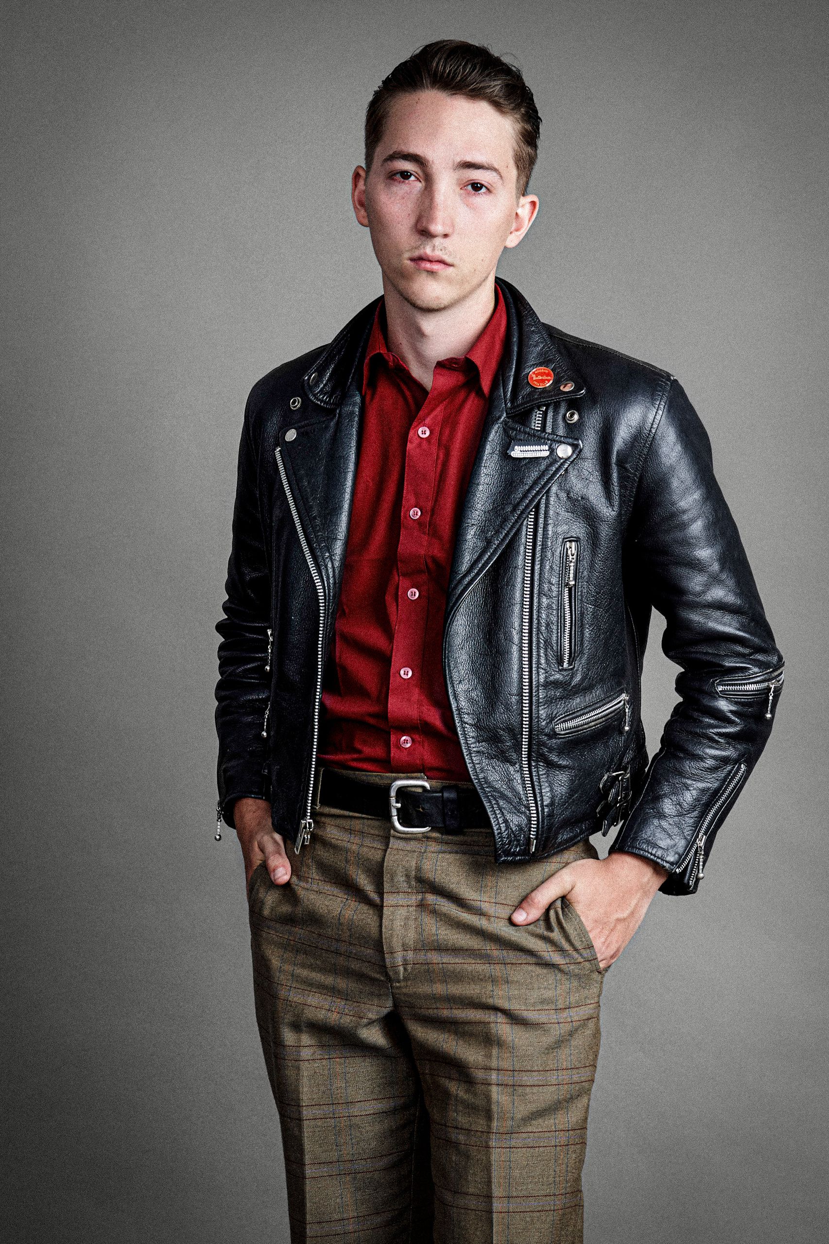 Model wearing a black leather jacket, red shirt and plaid pants in a fashion studio portrait.