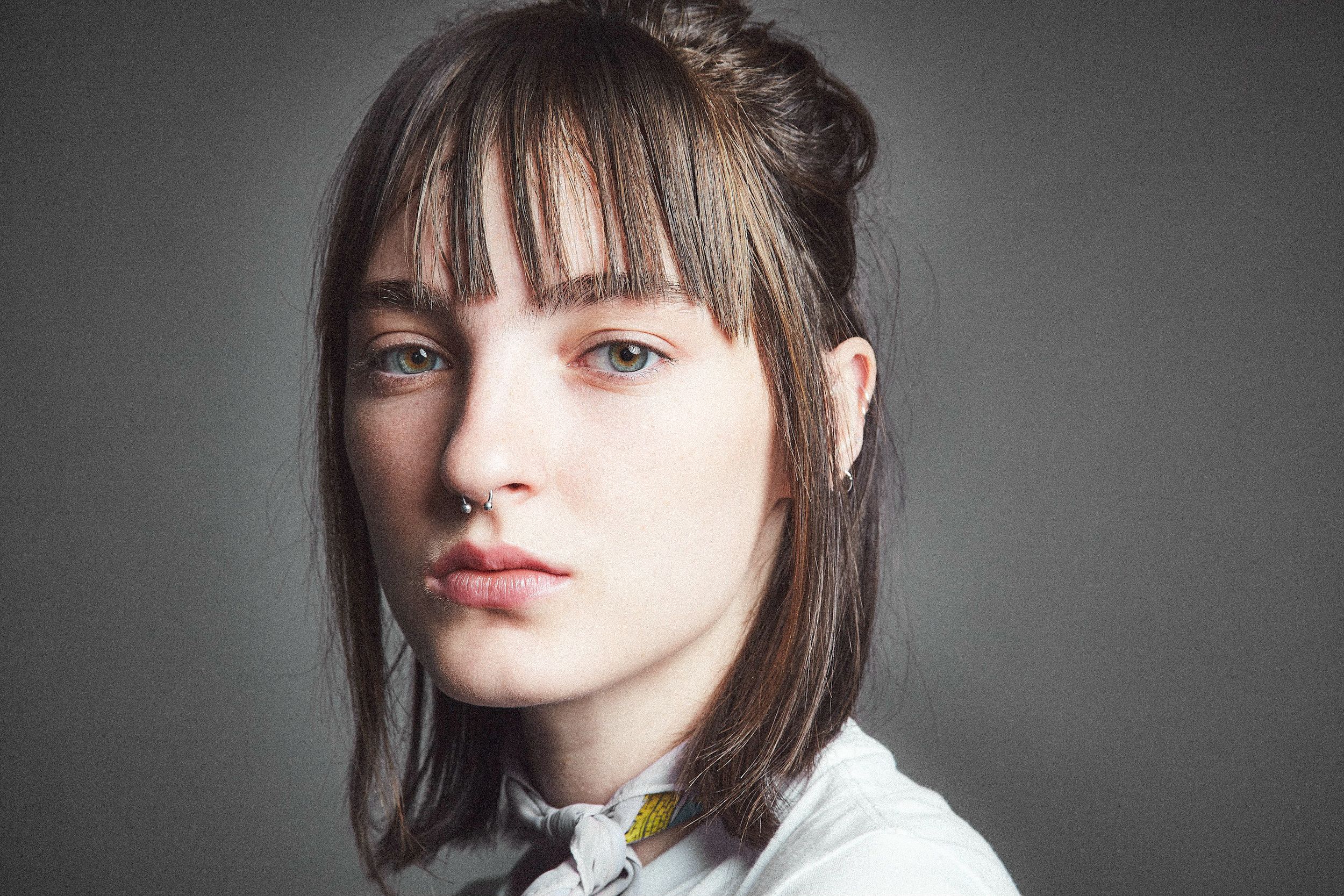 Portrait photo against gray background showing person with dark bangs and white collared shirt in dramatic lighting.
