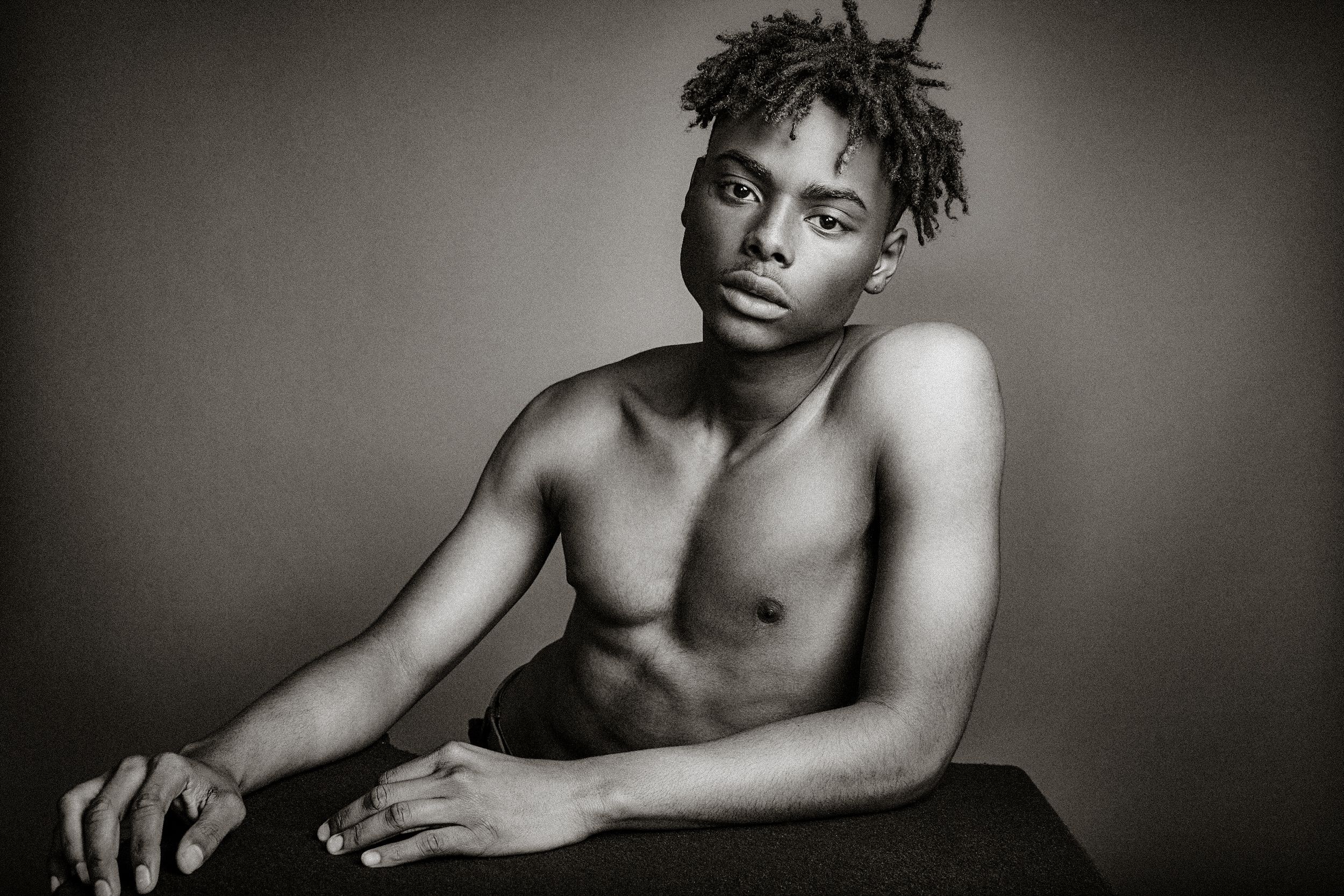 Artistic black and white portrait of a shirtless young person with dreadlocks against dark background.