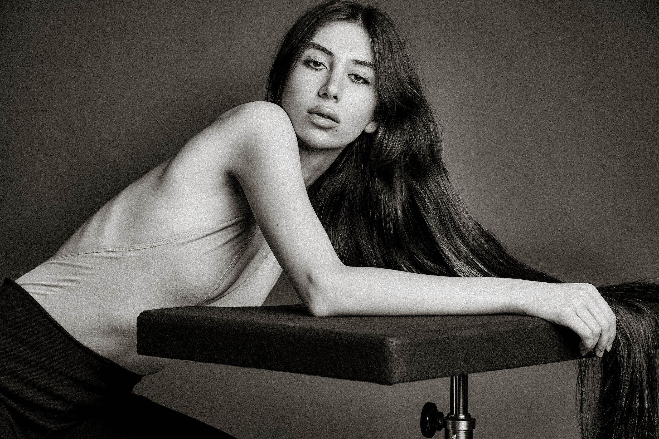 A black and white artistic portrait of a model with long dark hair reclining on a bench in a minimalist setting.