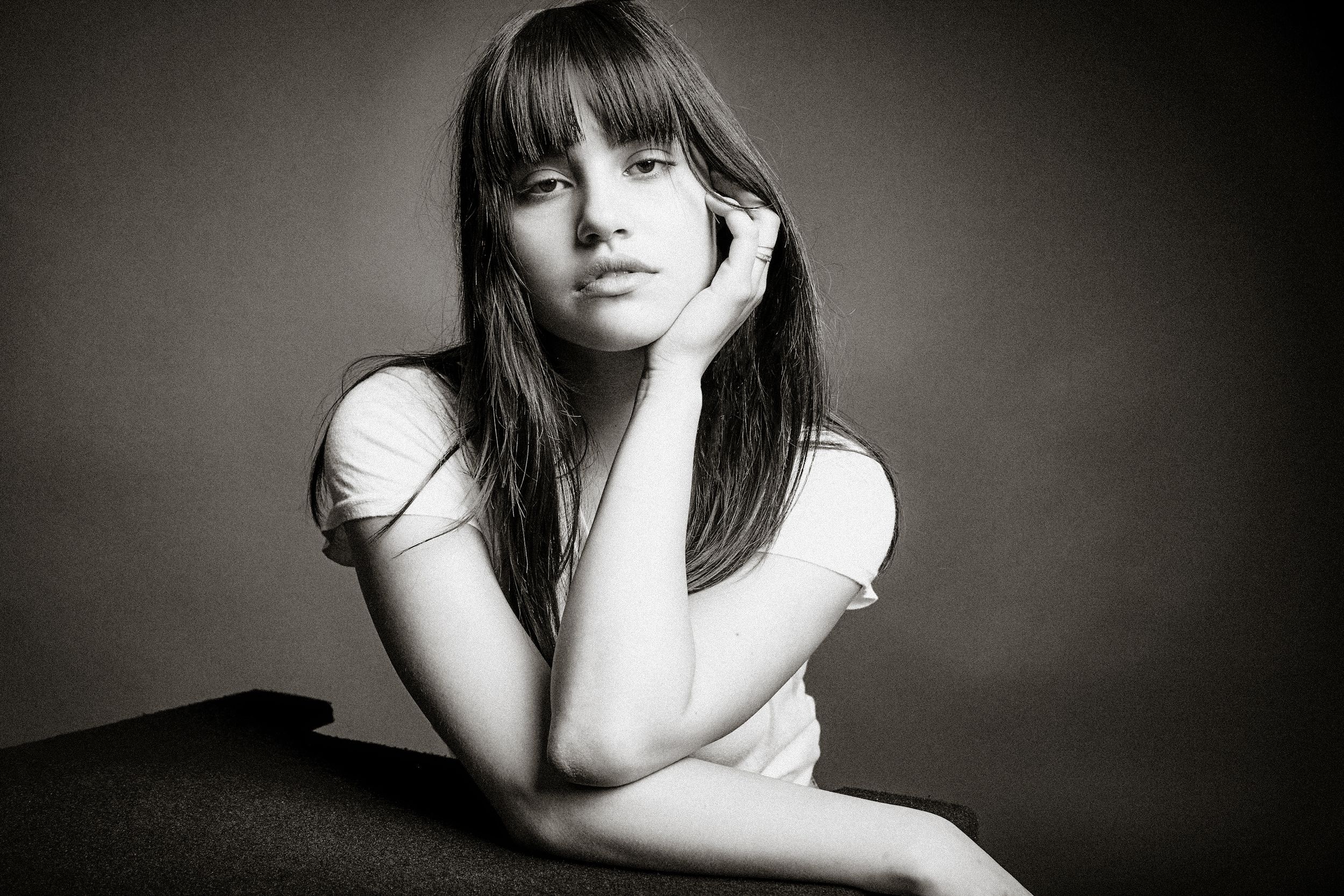 Black and white portrait of a person with long bangs and straight dark hair resting their chin on their hand.