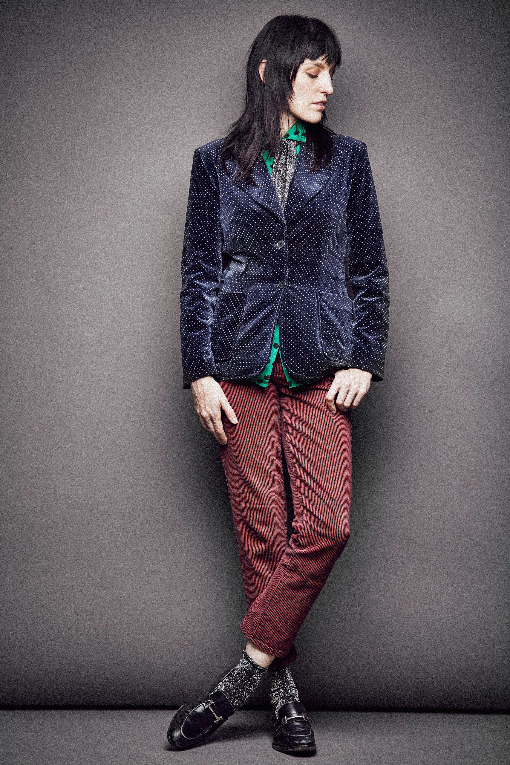 Fashion model wearing navy corduroy blazer with burgundy striped pants and black loafers against gray background.