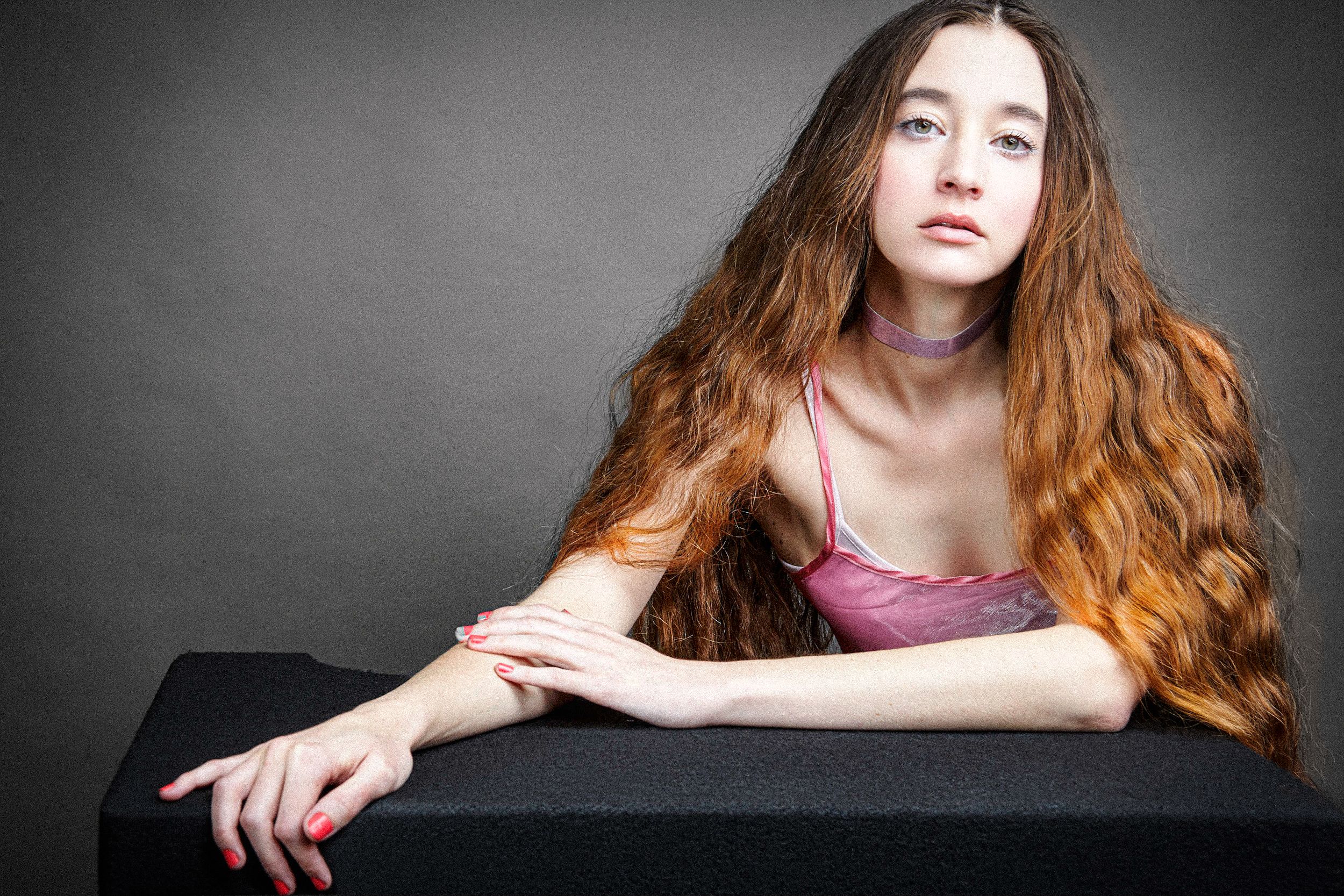 Portrait of someone with long wavy auburn hair wearing a pink top against a dark background.