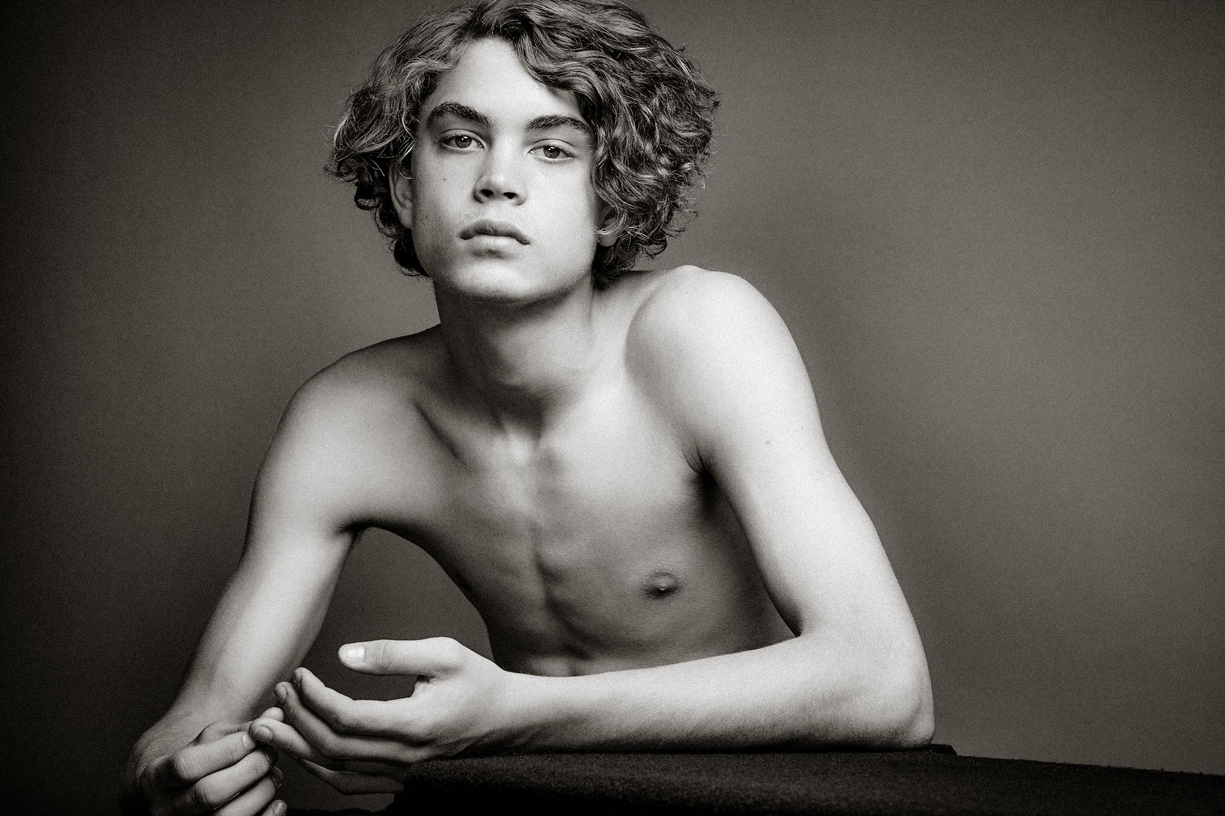 Black and white artistic portrait of a shirtless person with curly hair leaning on a table.