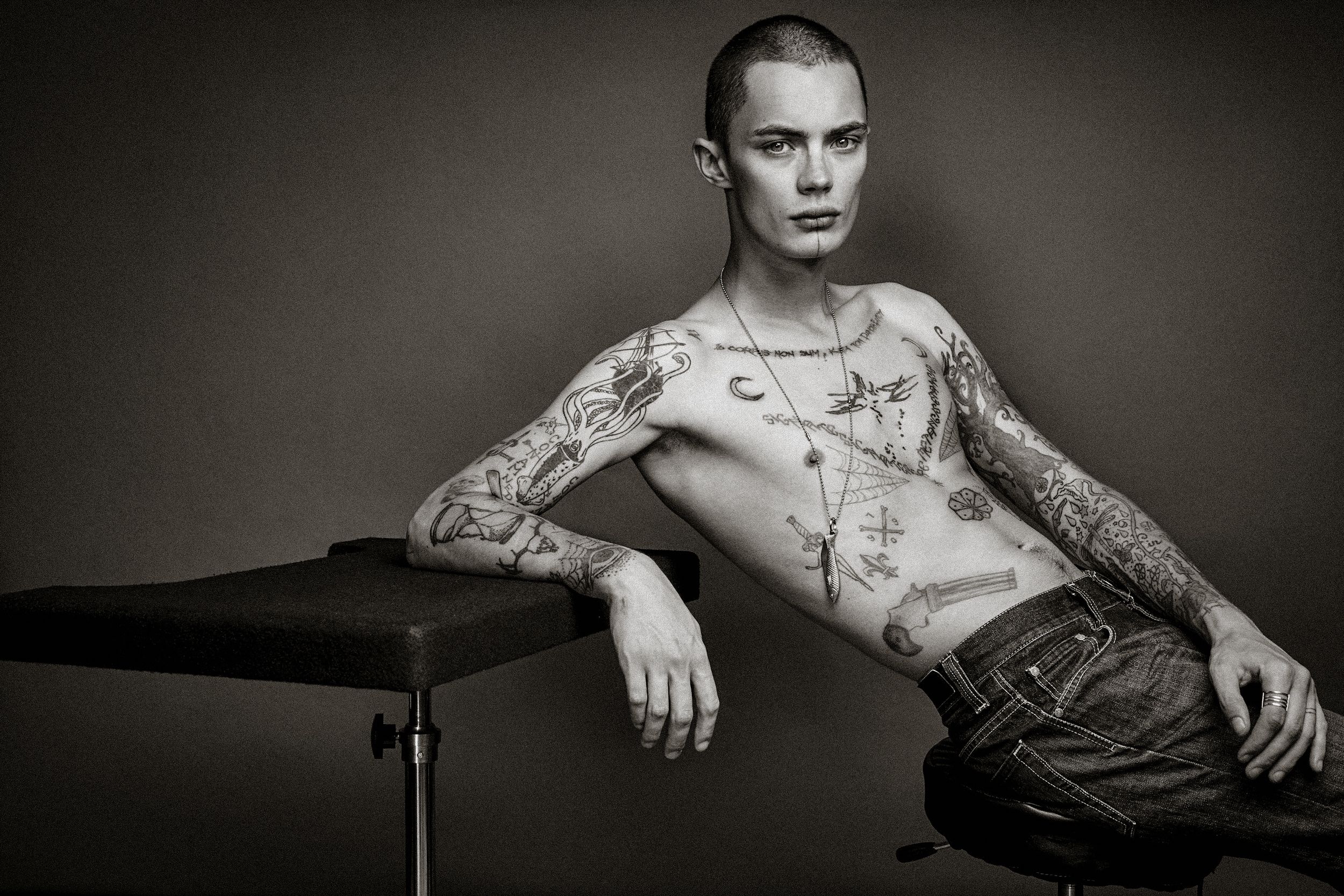 Black and white portrait of a shirtless person with tattoos sitting casually against a dark background.