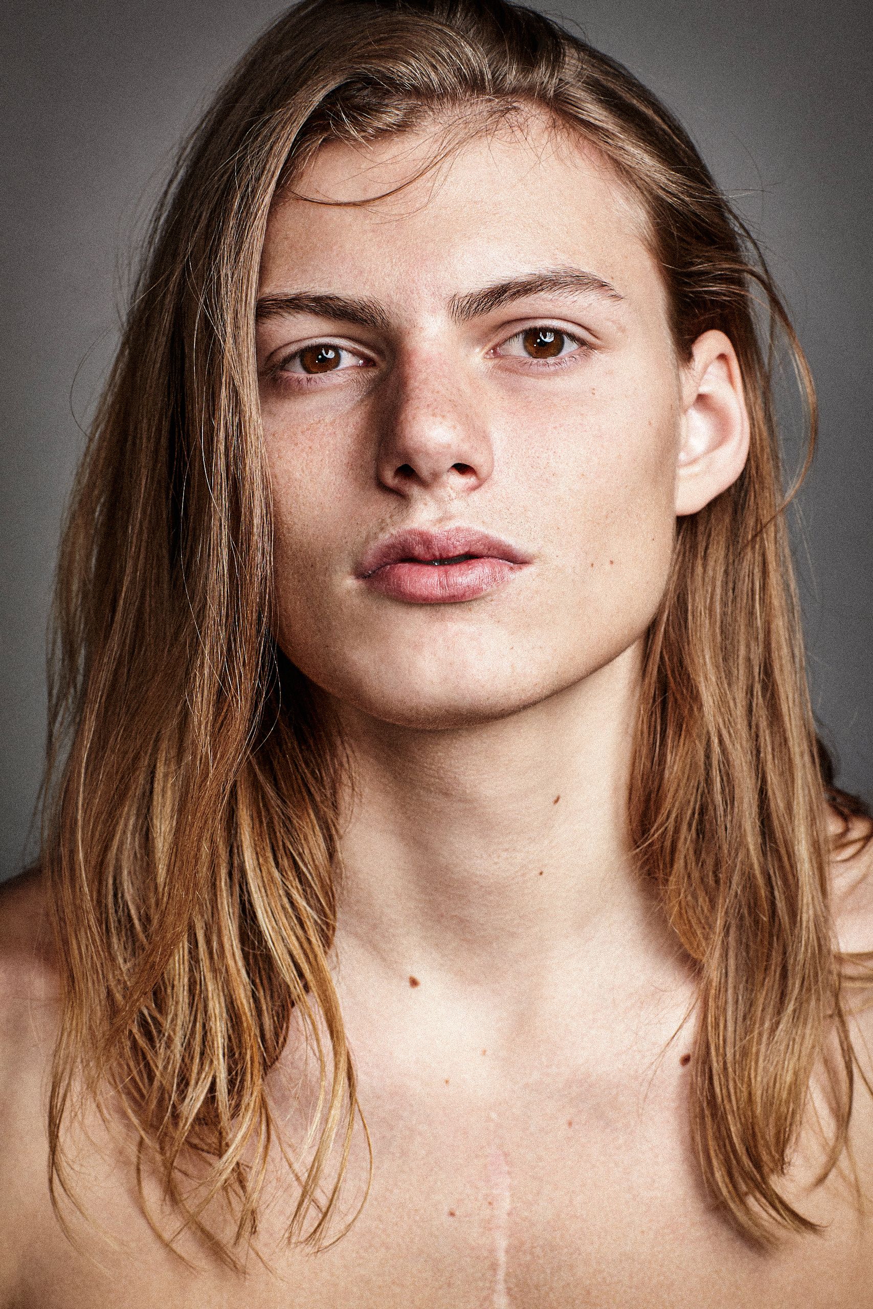 Close-up beauty portrait with natural lighting showing shoulder-length brown hair and minimal makeup against grey backdrop.