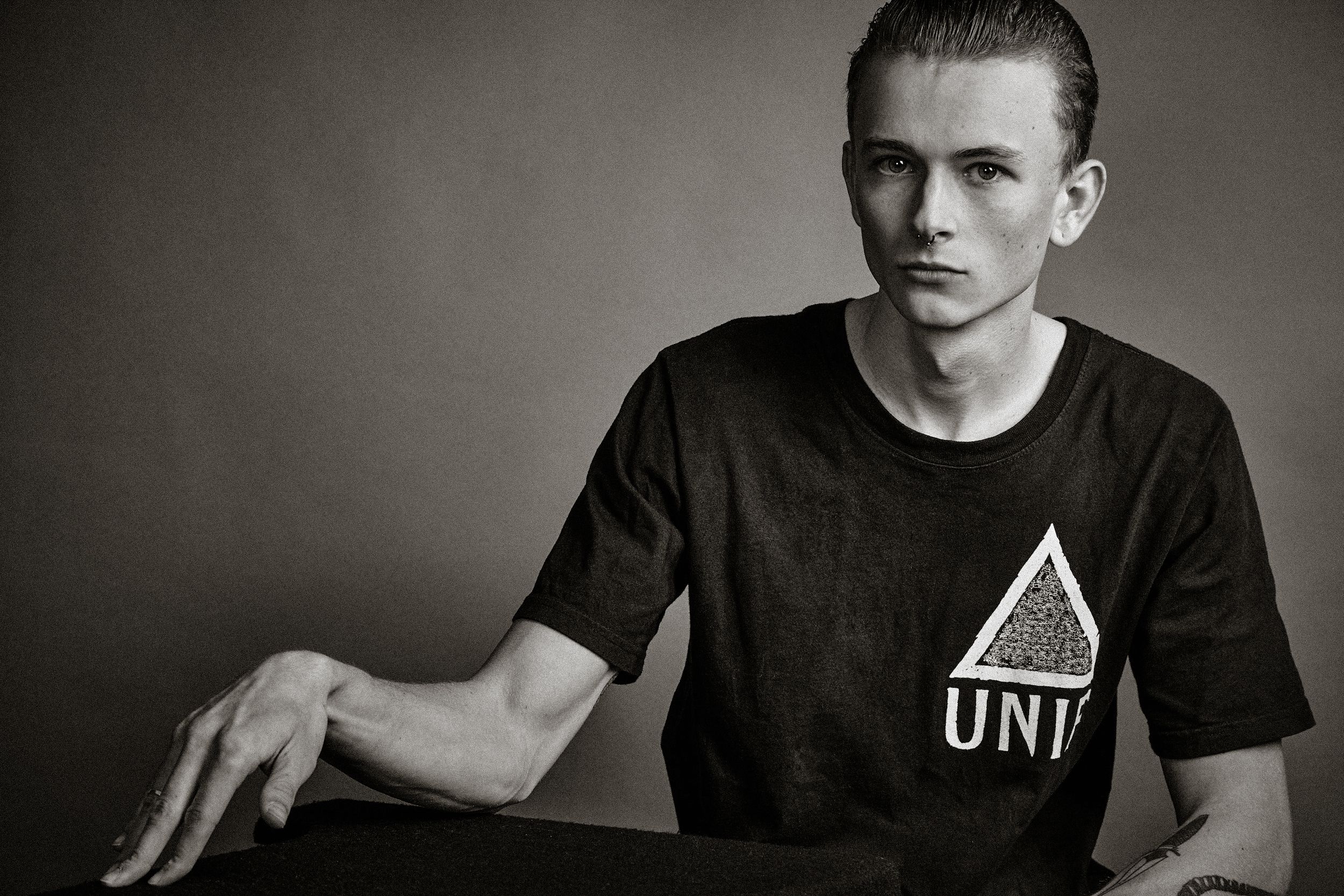 Black and white studio portrait of a person wearing a dark t-shirt against a gradient background.