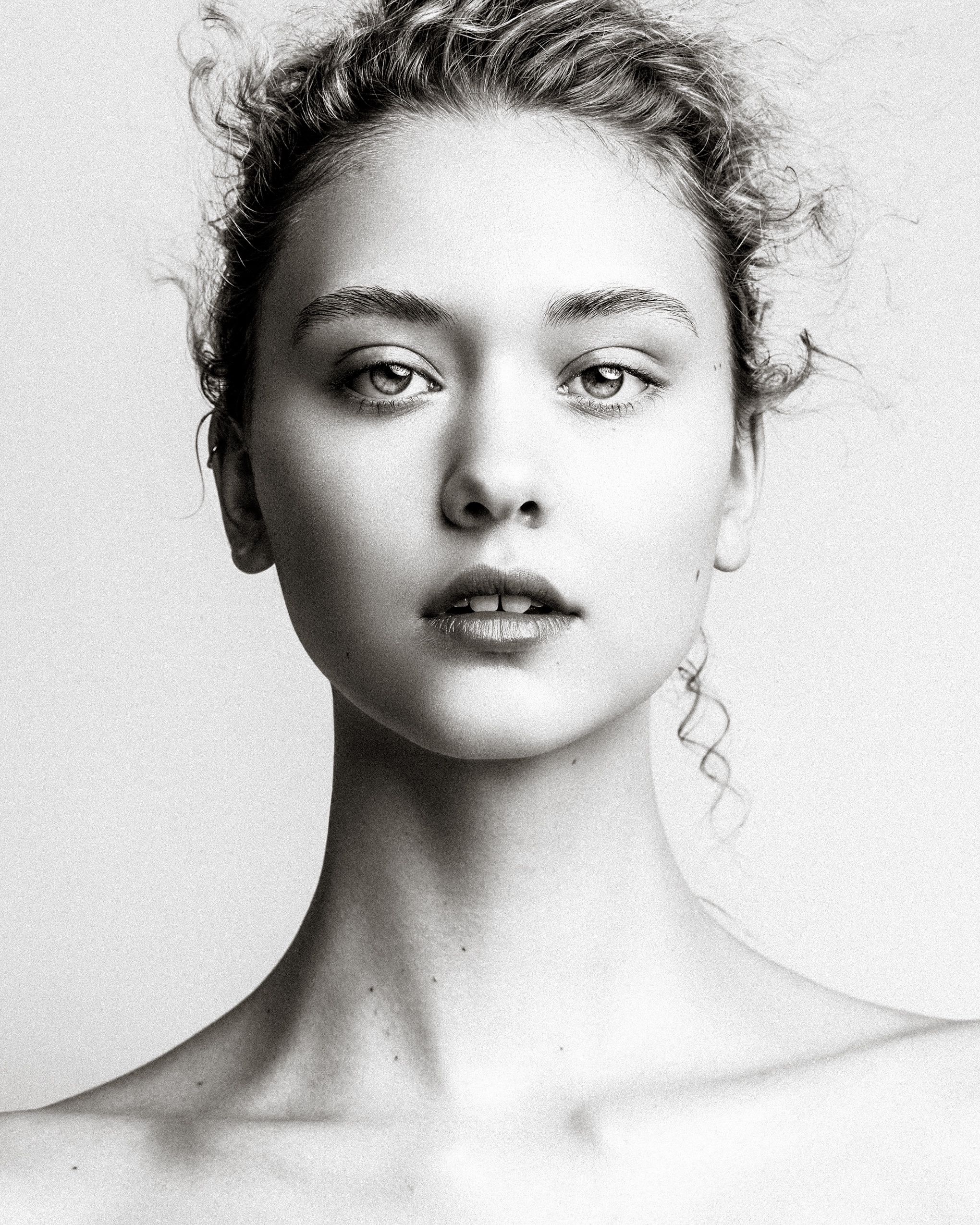 Black and white portrait photograph showing dramatic lighting and natural styling with wispy hair and bare shoulders.