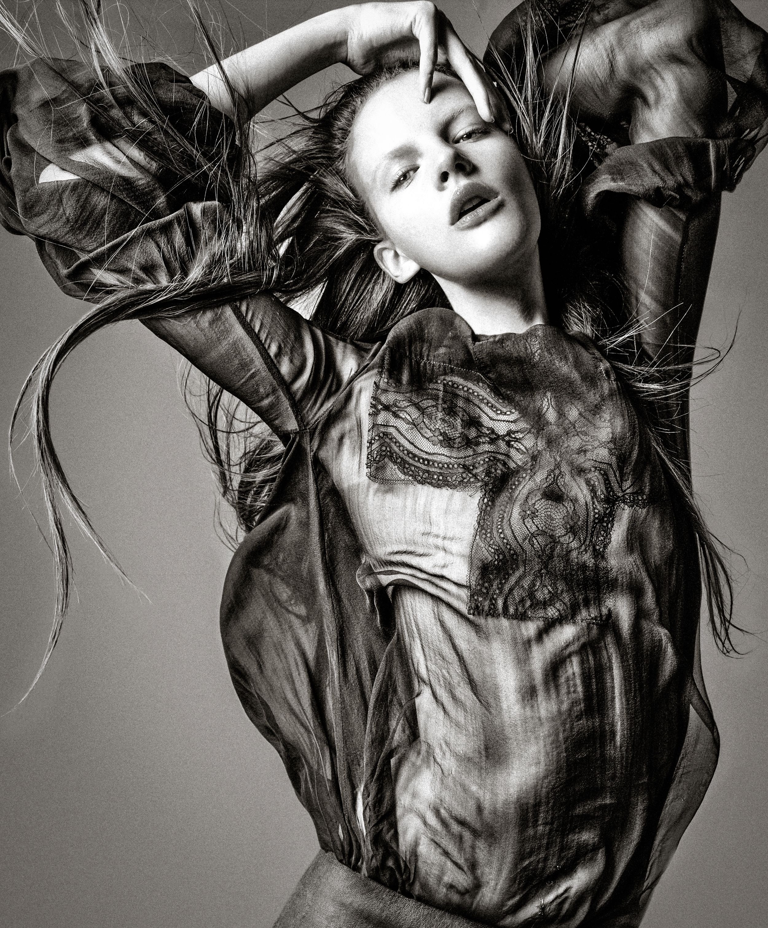 Dynamic black and white fashion photograph with flowing hair and fabric creating dramatic movement against a neutral background.