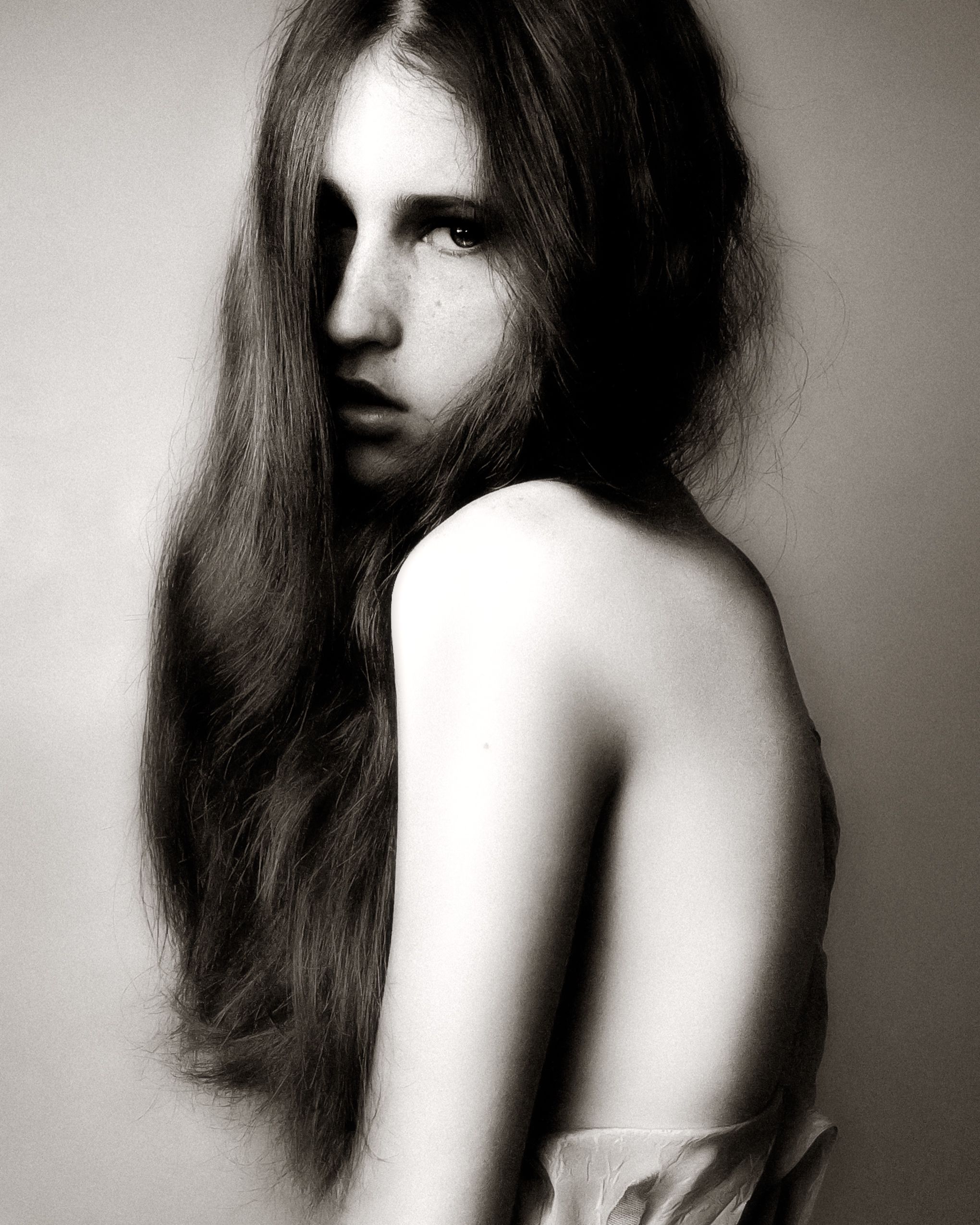 Black and white artistic portrait of a person with long dark hair looking over their bare shoulder in dramatic lighting.