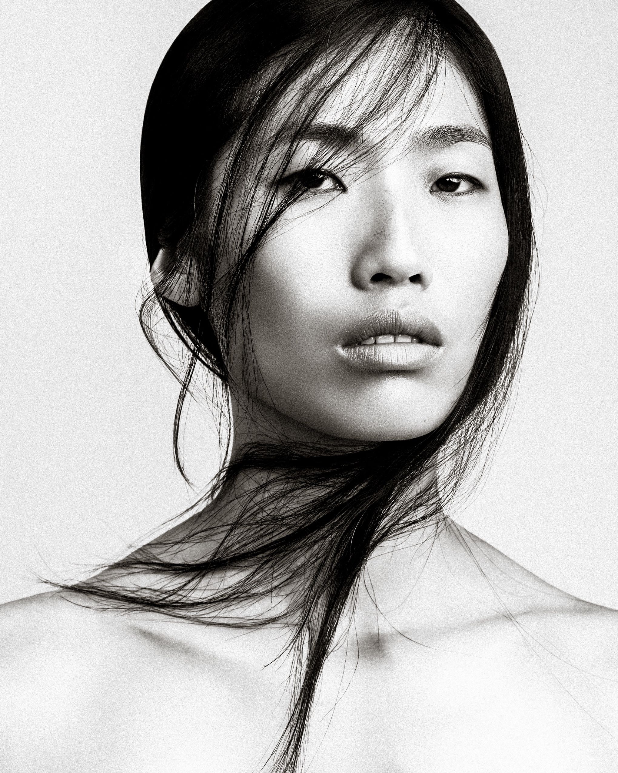 Black and white artistic portrait with dramatic lighting showing wind-swept hair and natural makeup against white background.