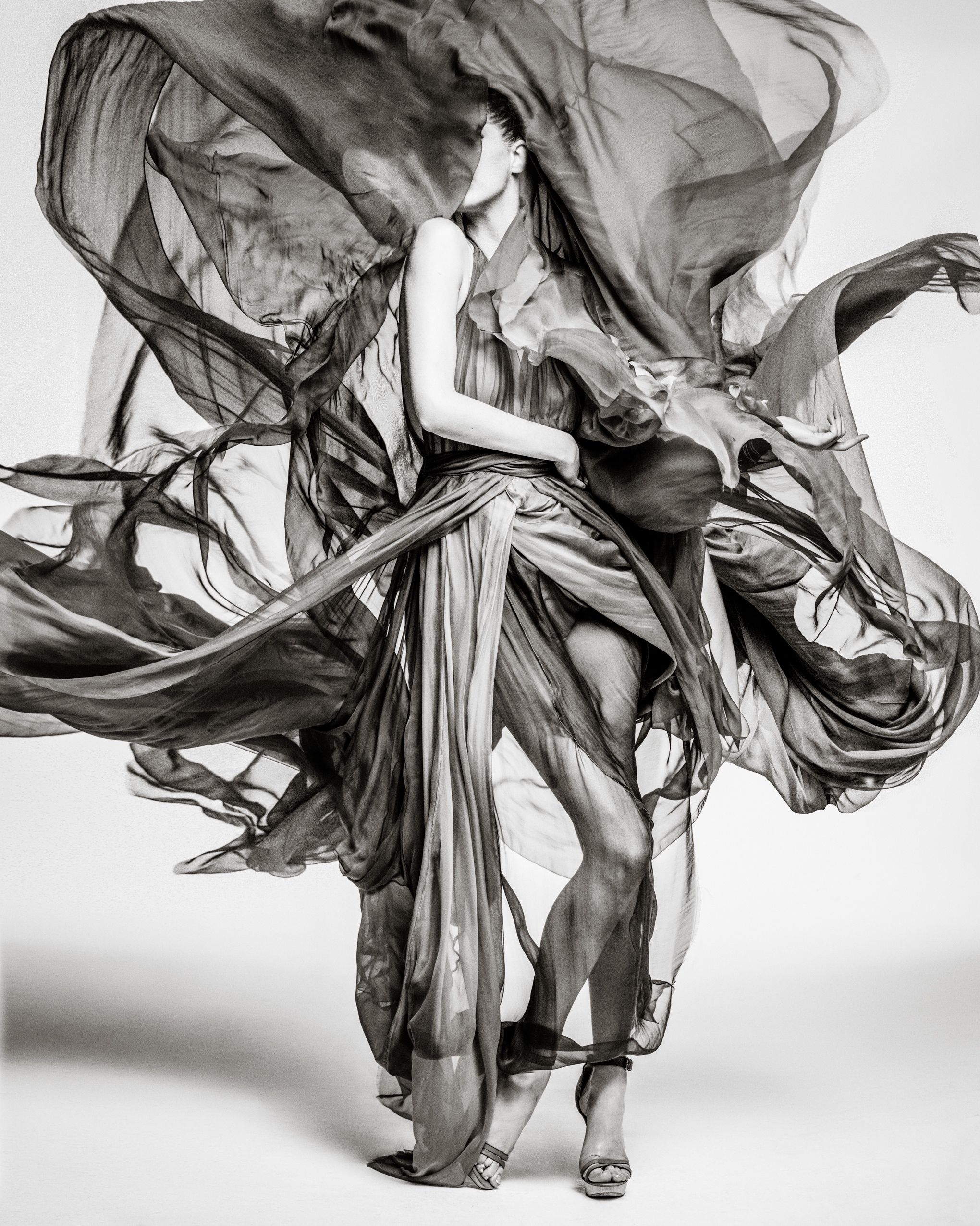 Black and white artistic fashion photograph showing flowing fabric dramatically billowing around a figure in motion.