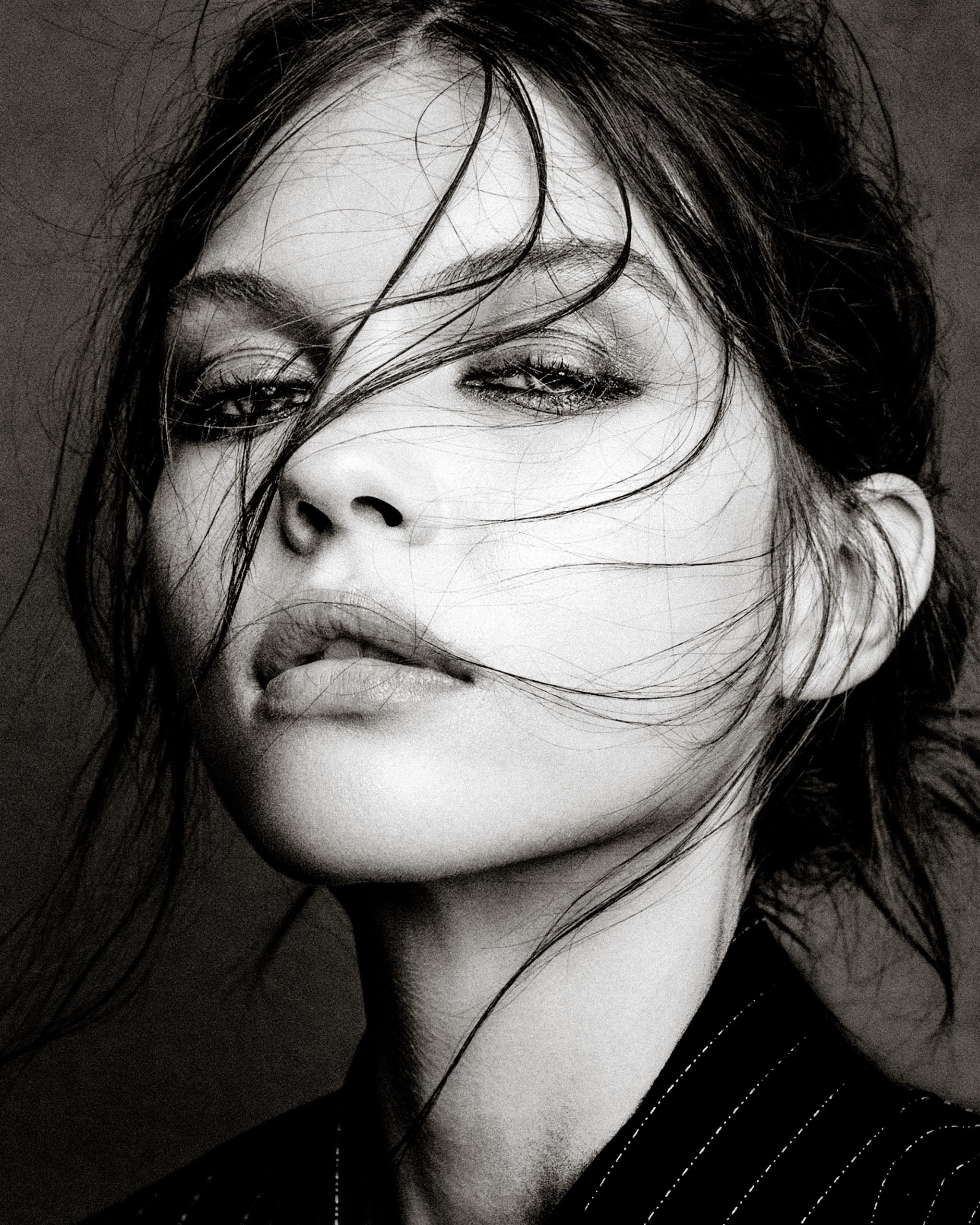 Dramatic black and white portrait with windswept hair against dark background showing intense gaze and defined features.