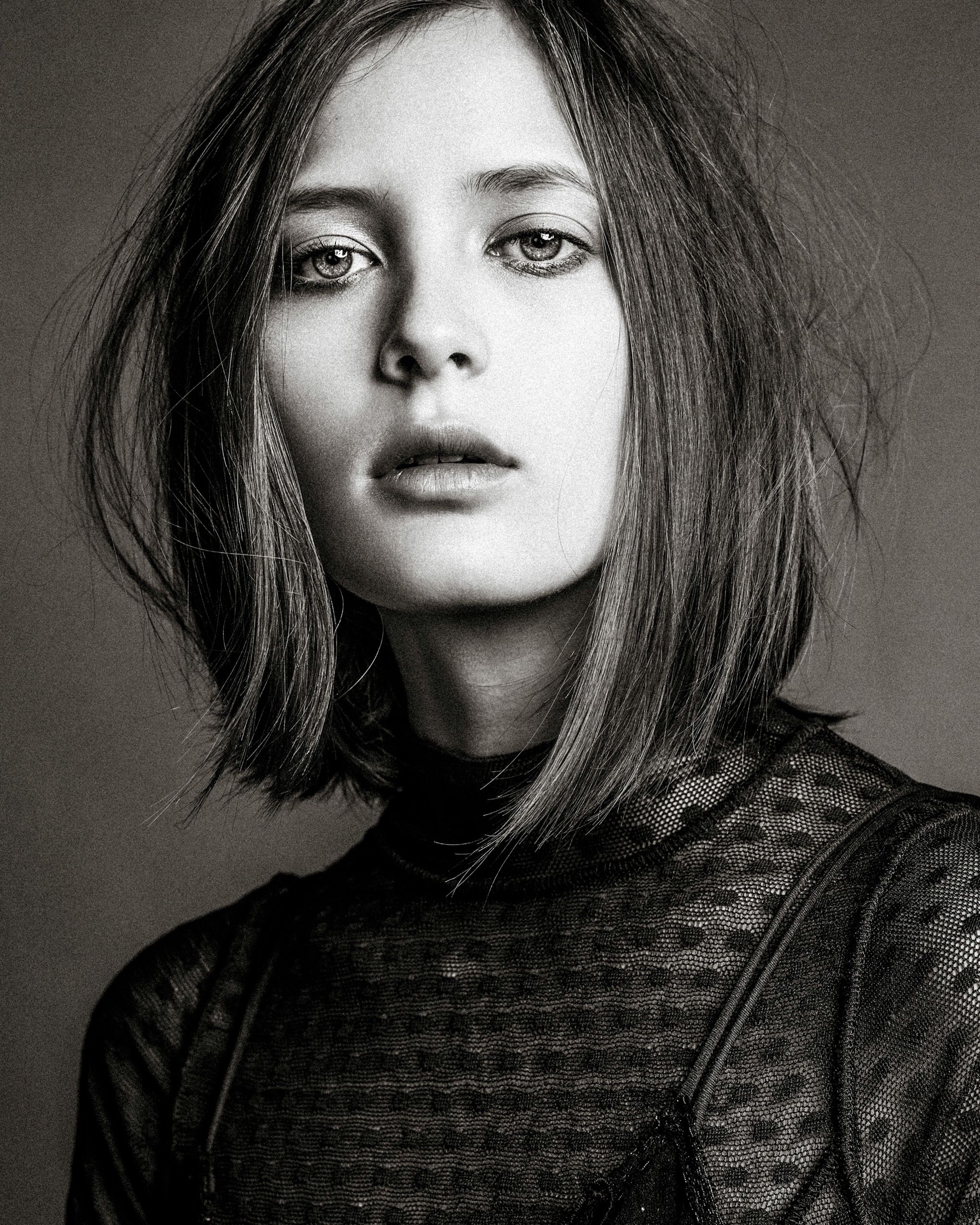 Black and white portrait photograph showing tousled hair and wearing a dark patterned sweater against a gray backdrop.