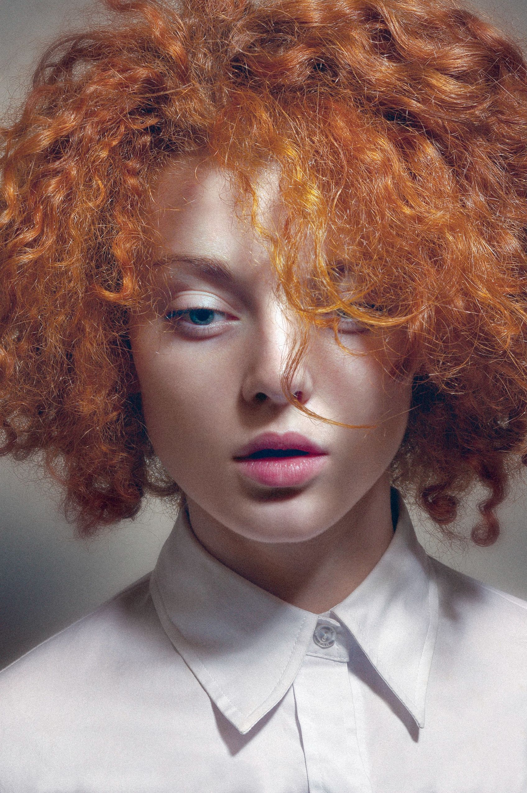 Fashion portrait with vibrant orange curly hair and minimal makeup against white background.