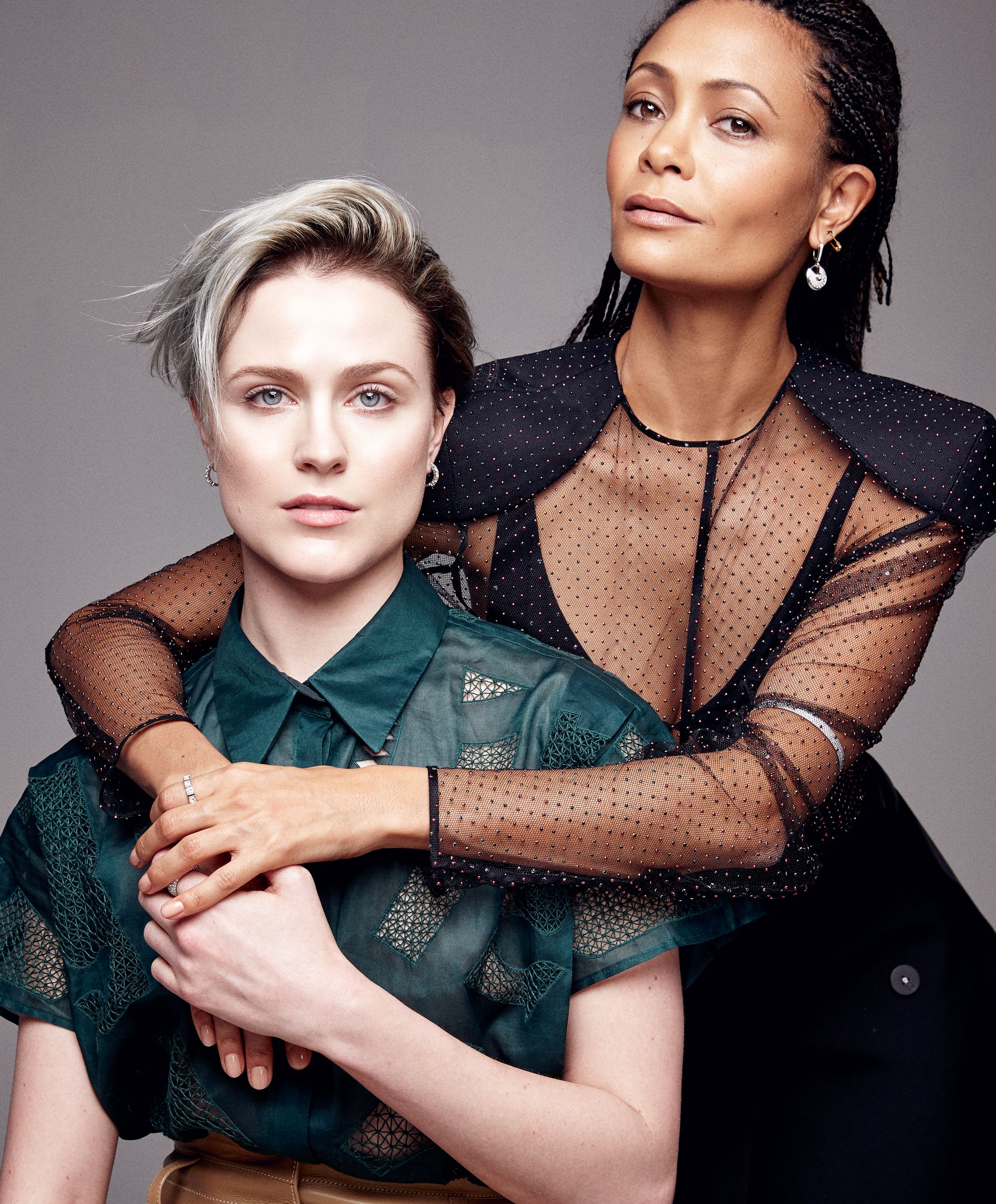 Two models pose together in a fashion portrait wearing dark sheer and leather outfits against a gray background.