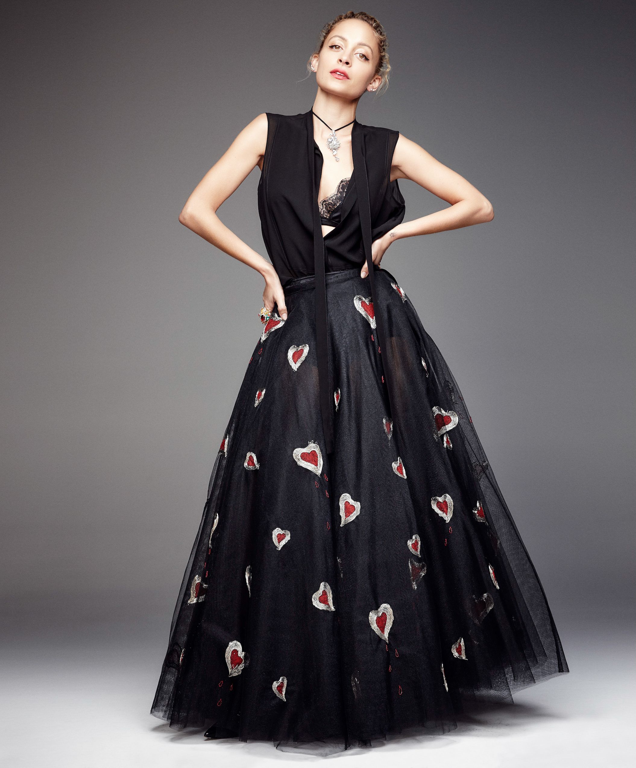 A model poses in a dramatic black tulle gown adorned with red heart appliques against a gray studio backdrop.