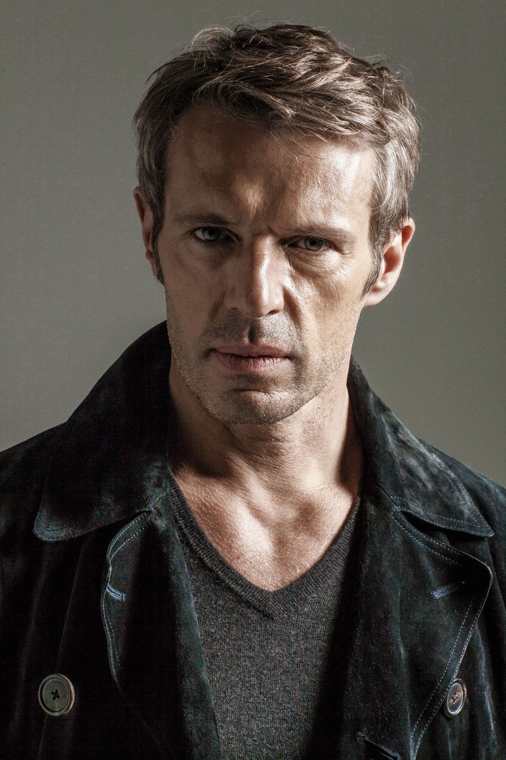 Dramatic studio portrait of someone wearing a dark jacket and gray v-neck shirt against neutral background.