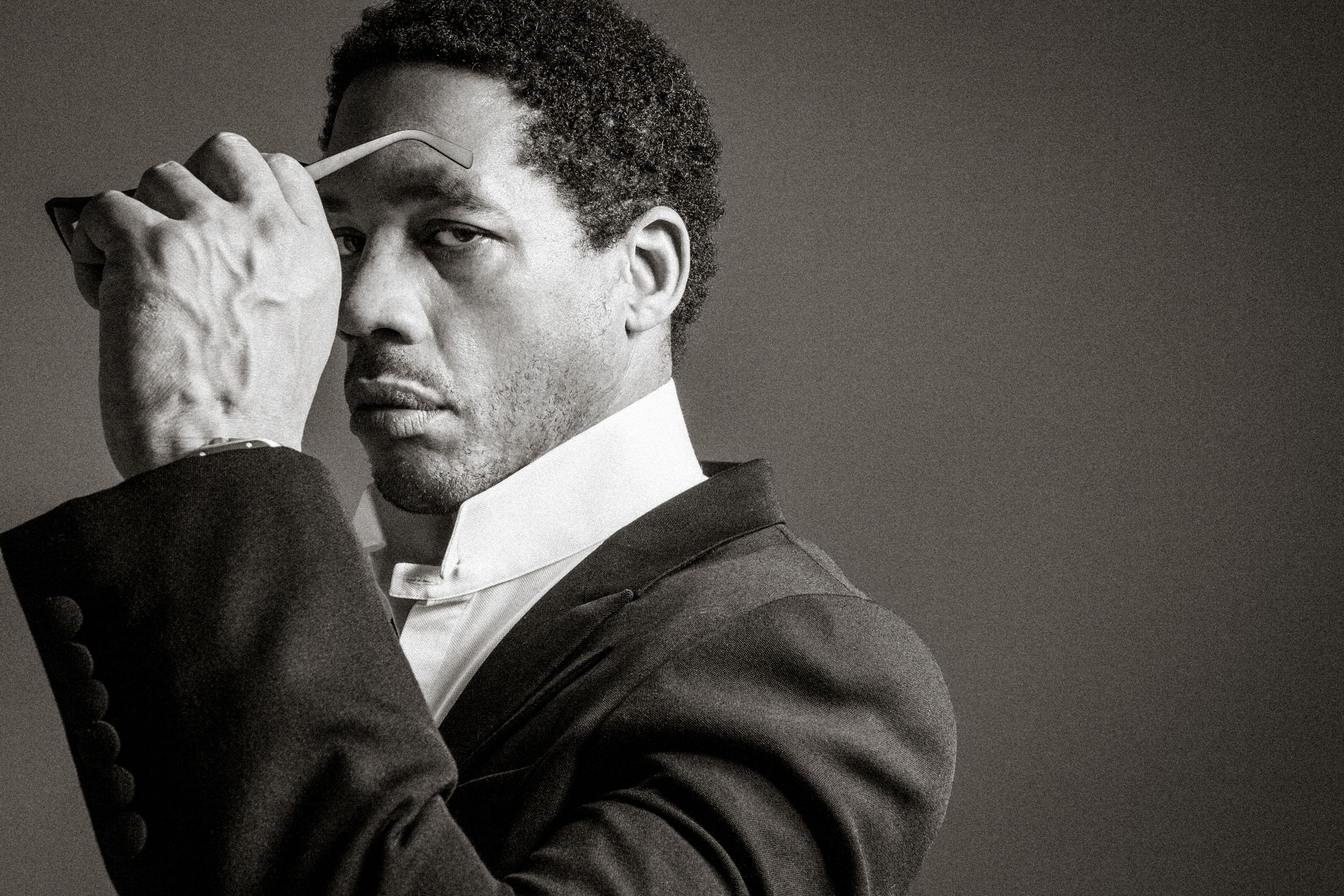 Black and white portrait of a person in a suit with hand raised to forehead in contemplative pose against gray background.